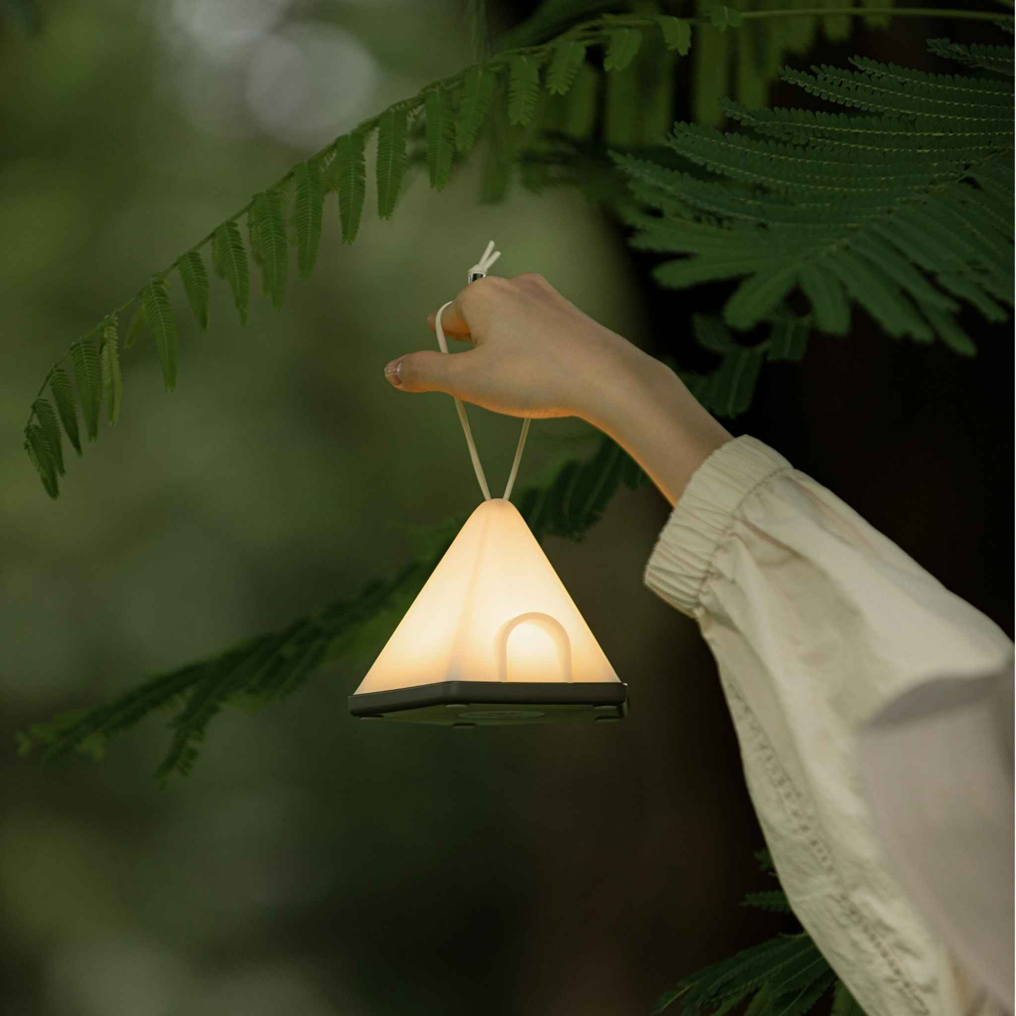 Eine Hand, die eine beleuchtete, weiße, dreieckige Lampe in einer Waldumgebung mit grünem Laub hält.