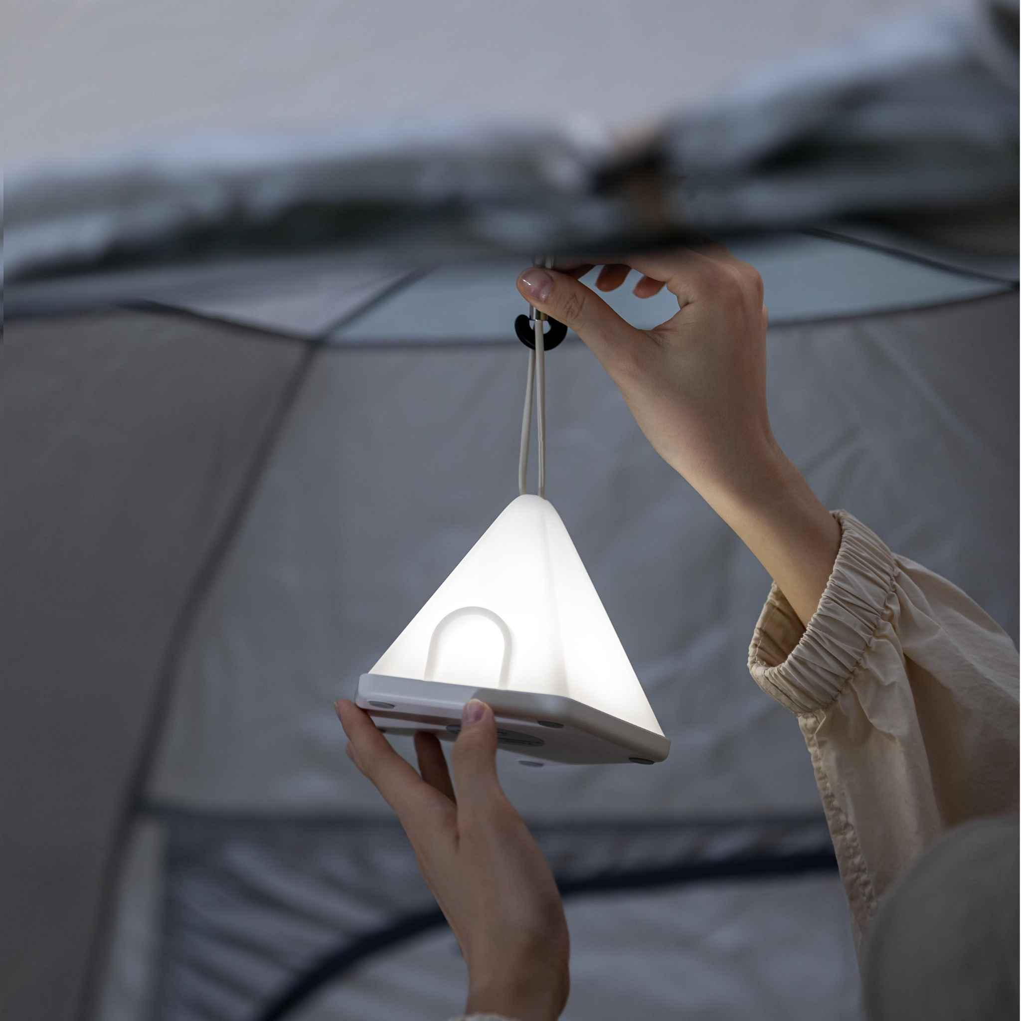 Hände halten eine leuchtende, weiße, dreieckige Lampe in einem Zelt.