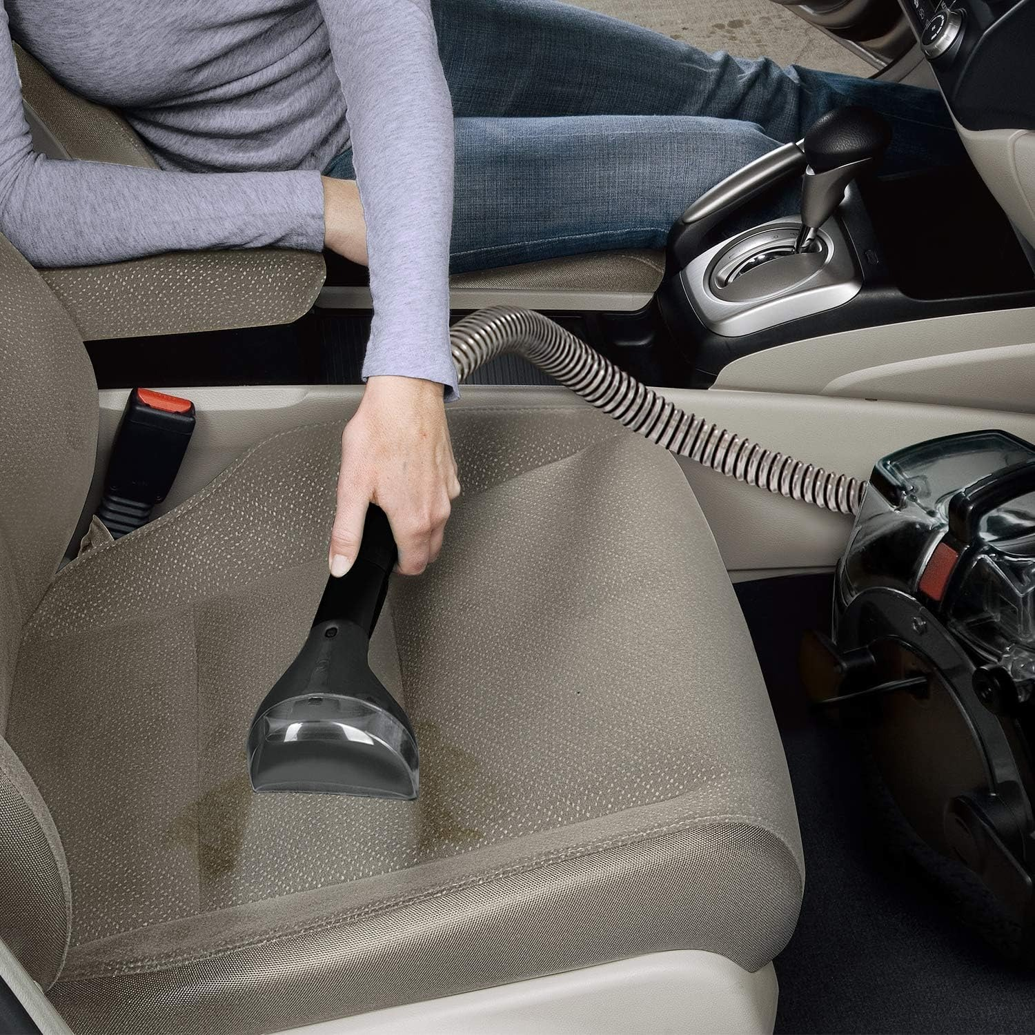 Una persona sta usando un aspirapolvere su un sedile dell'auto. Il sedile è beige e l'aspirapolvere è nero.