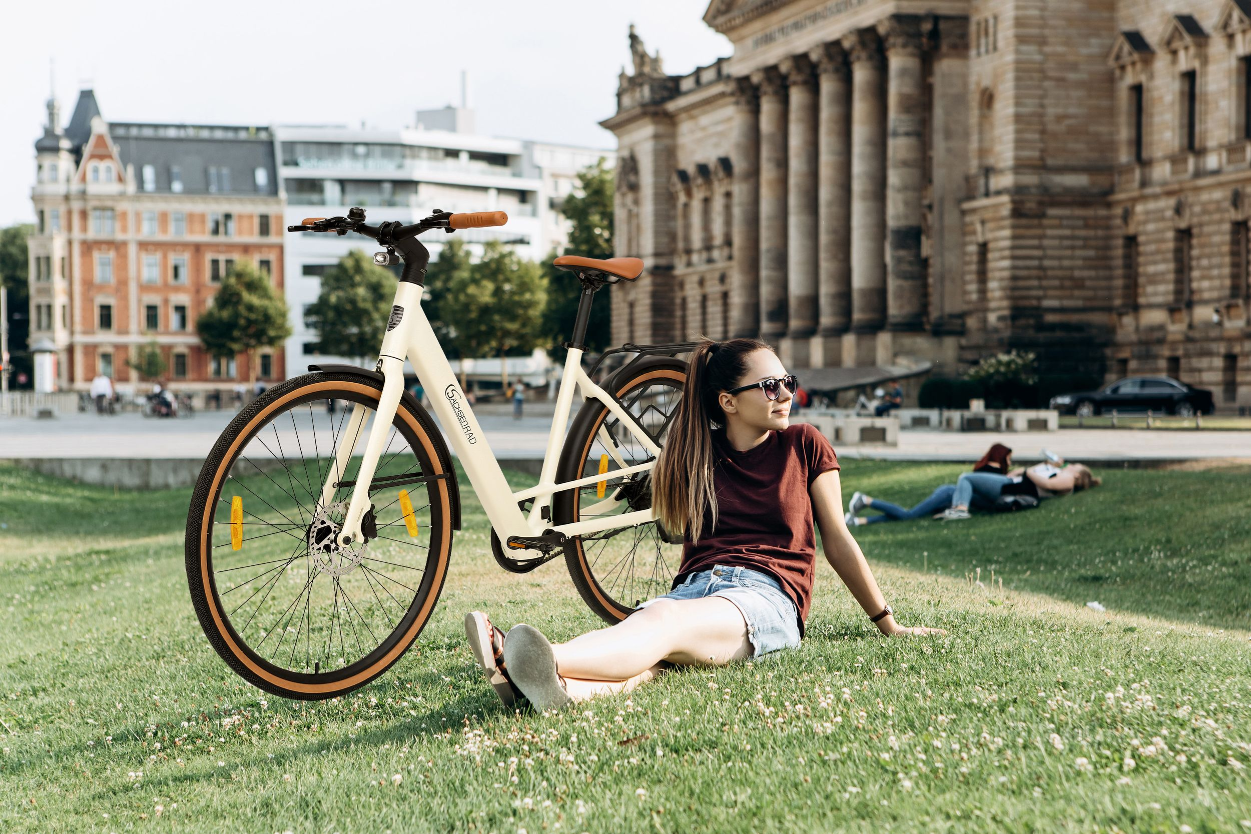 Frau sitzt auf Gras neben weißem Fahrrad, mit Stadt im Hintergrund.
