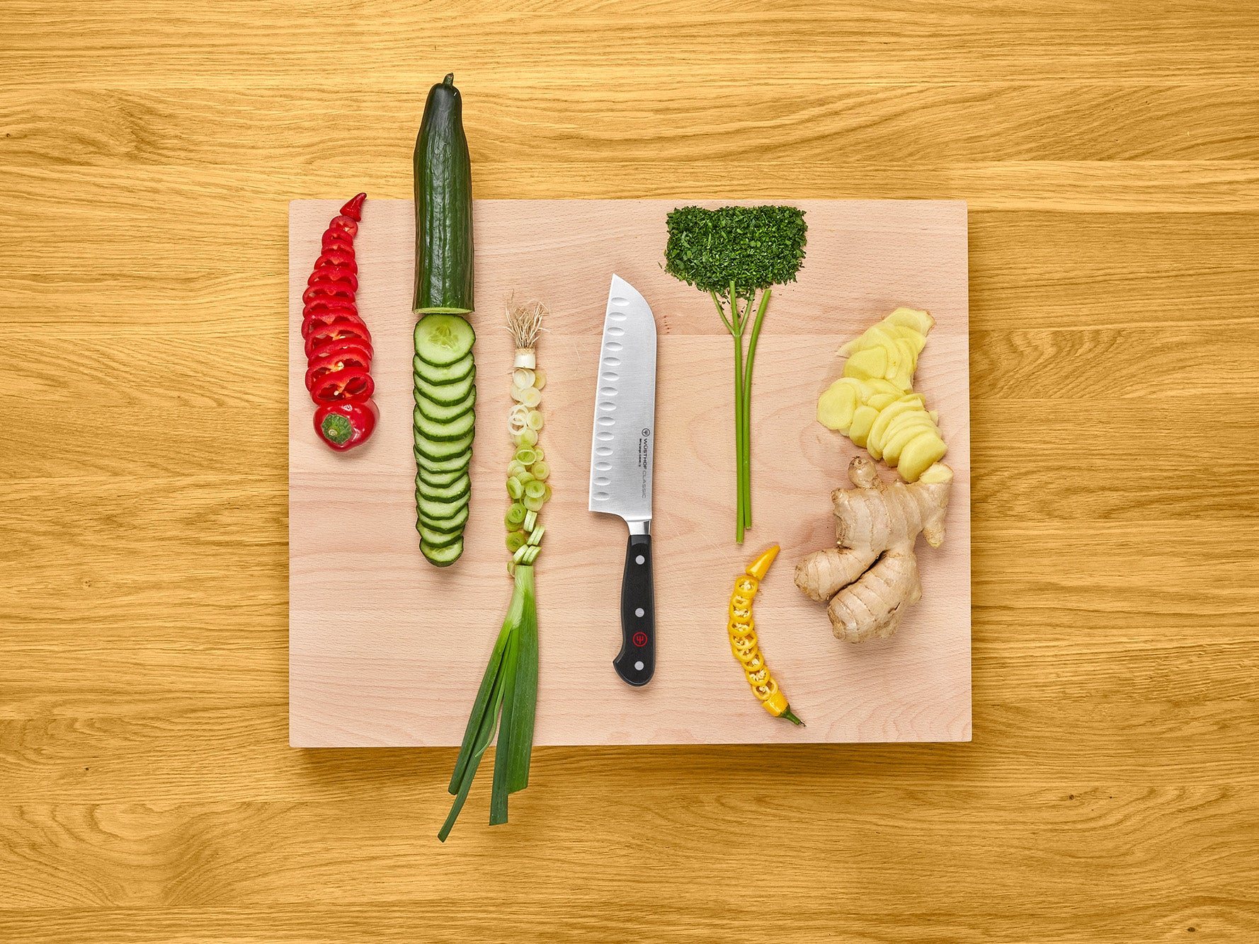 Una tabla de cortar de madera con verduras dispuestas: pimiento rojo, pepino, cebolletas, perejil, jengibre y pimiento amarillo.