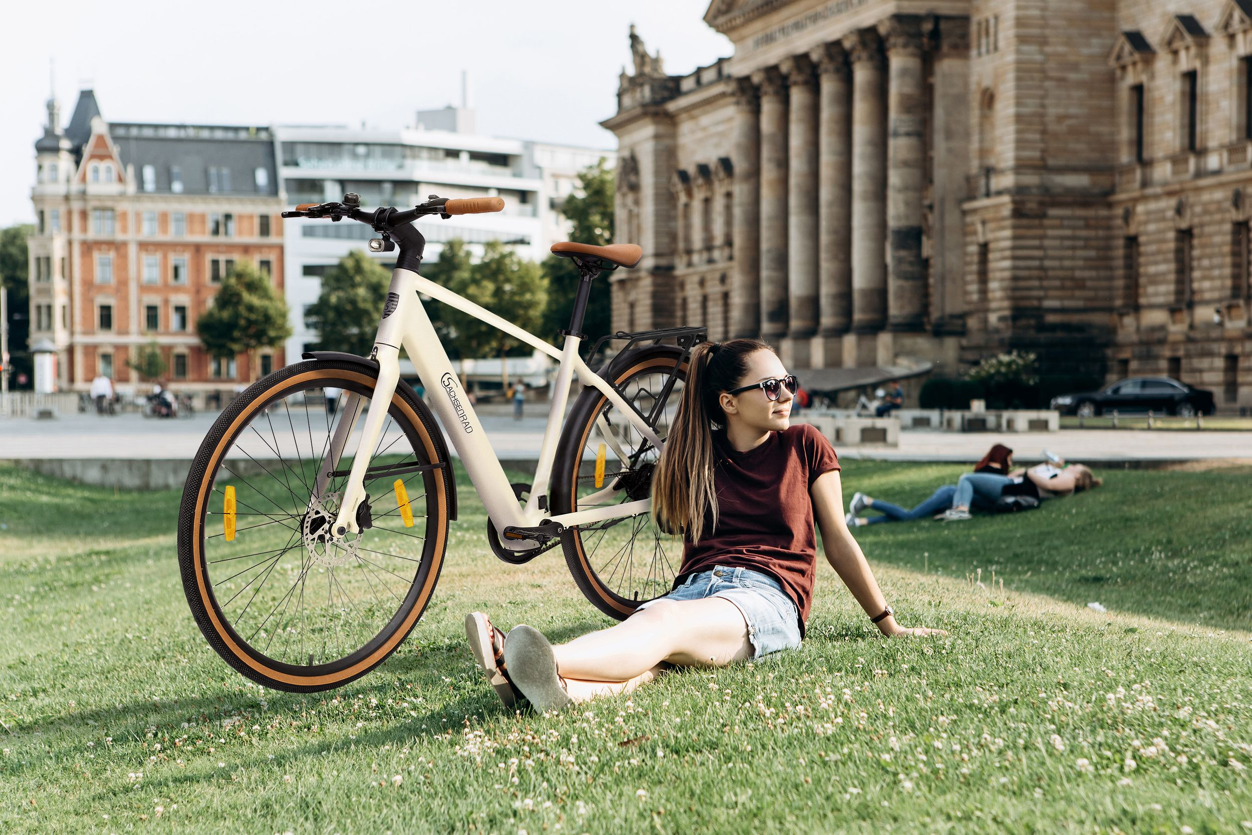Eine Frau sitzt auf Gras neben einem cremefarbenen Fahrrad. Ein Gebäude ist im Hintergrund.