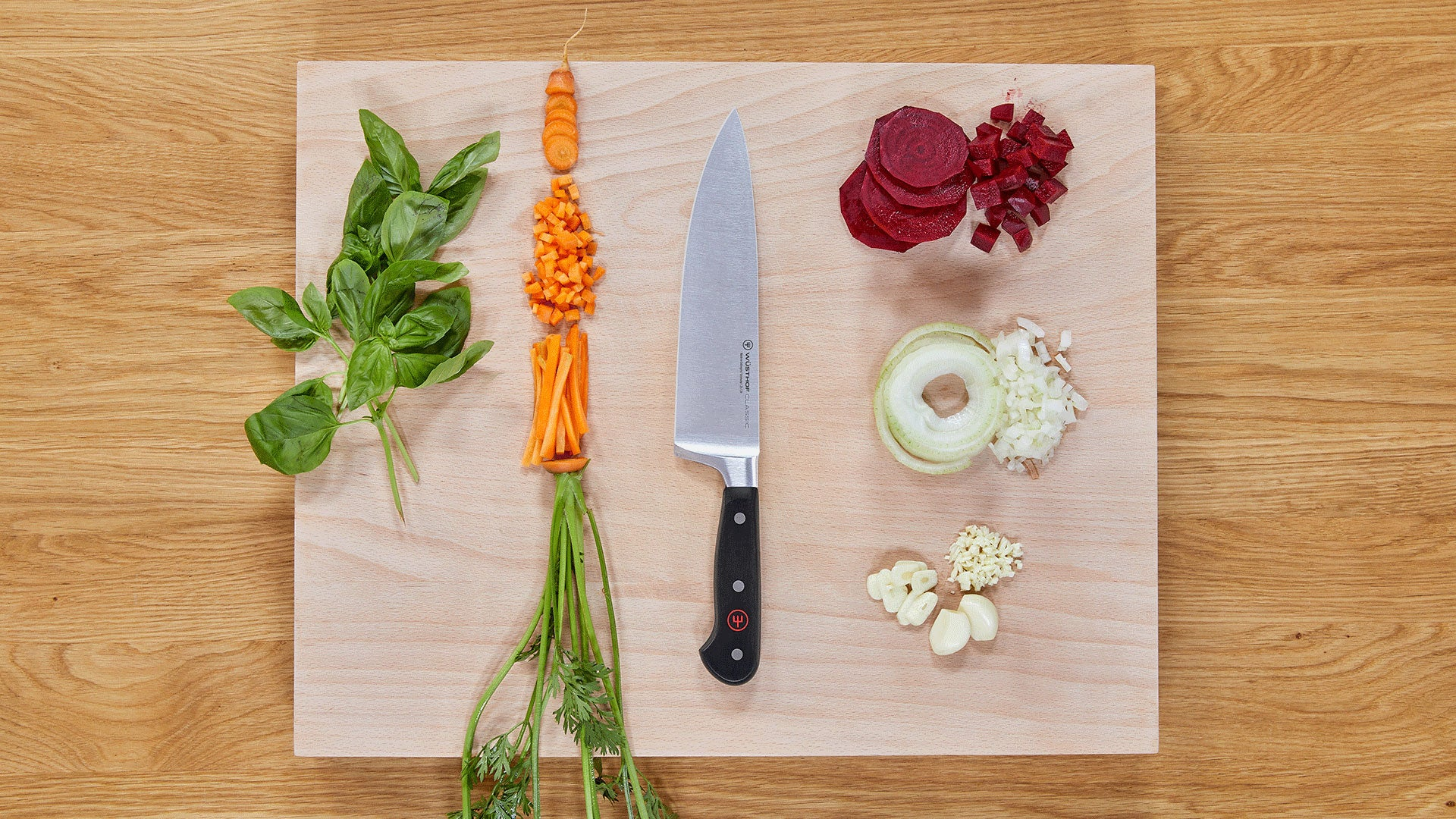 Una tabla de cortar de madera con un cuchillo y verduras dispuestas: albahaca, zanahorias, remolacha, cebolla, ajo y zanahorias.