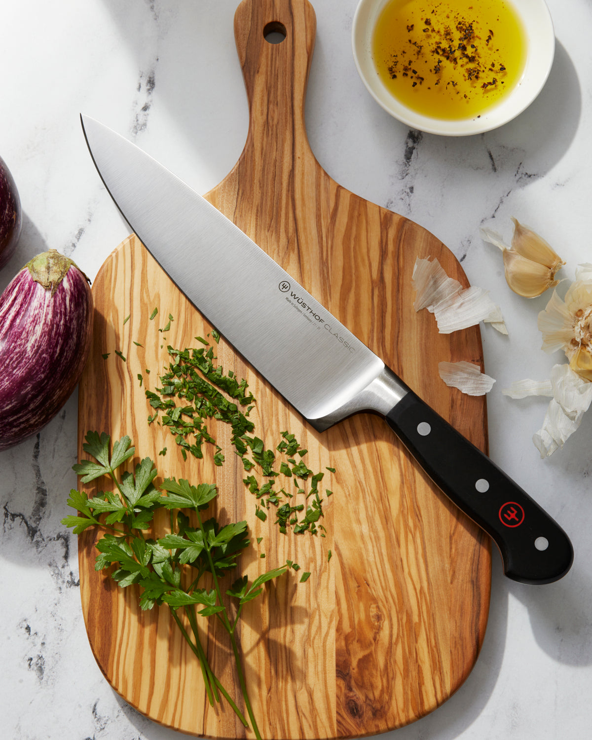 Un cuchillo de cocina grande sobre una tabla de cortar de madera, con perejil, aceite y ajo cerca.