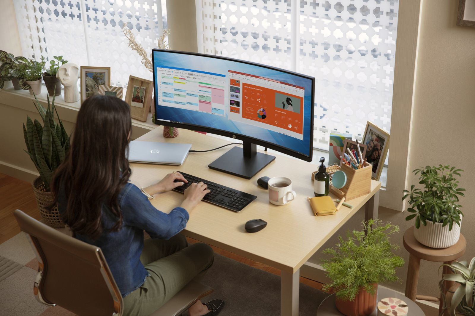 Une femme assise à un bureau avec un moniteur HP incurvé affichant une feuille de calcul. Il y a un ordinateur portable, un clavier et d'autres objets.