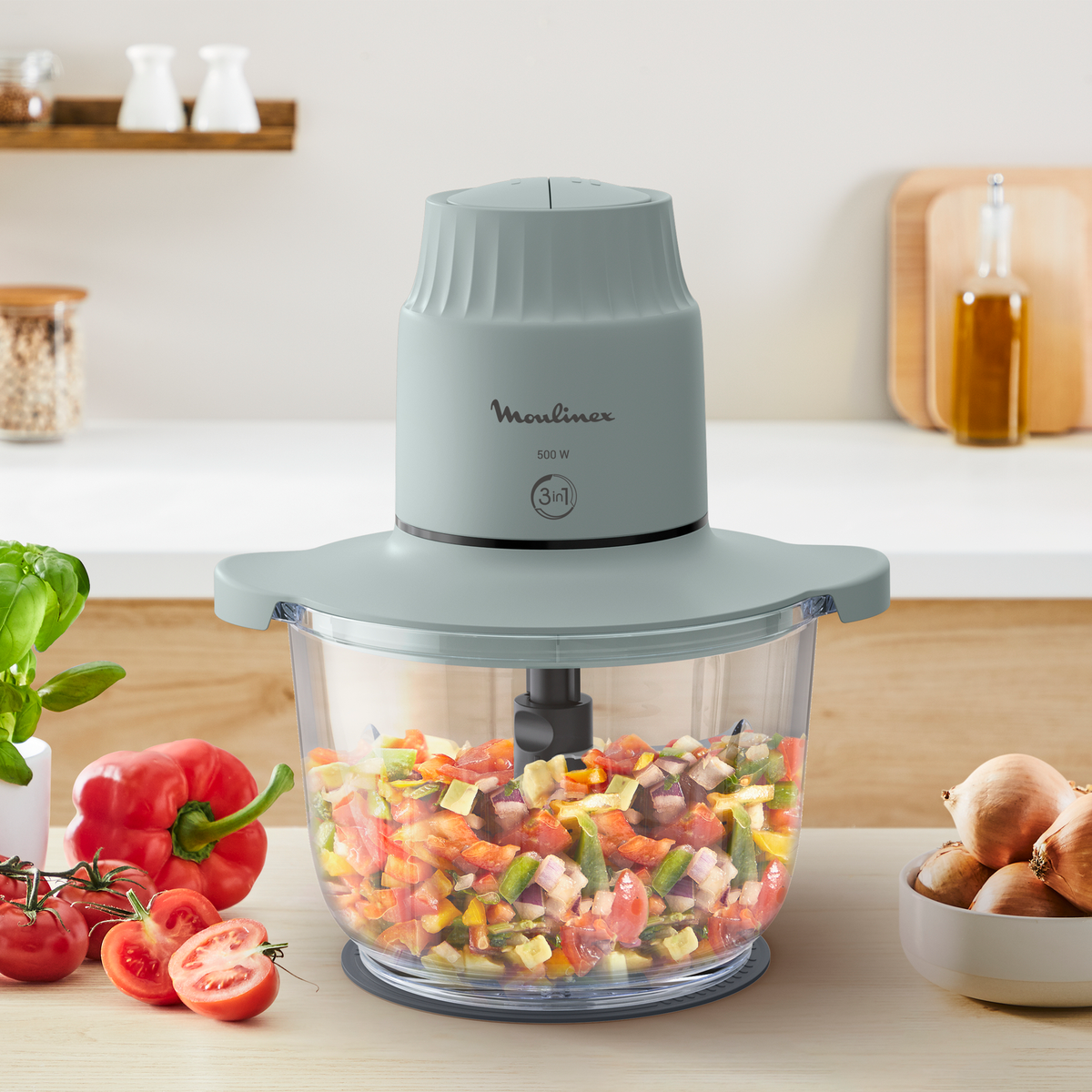 Un picador de alimentos Moulinex con verduras picadas. El fondo tiene tomates, cebollas y albahaca.
