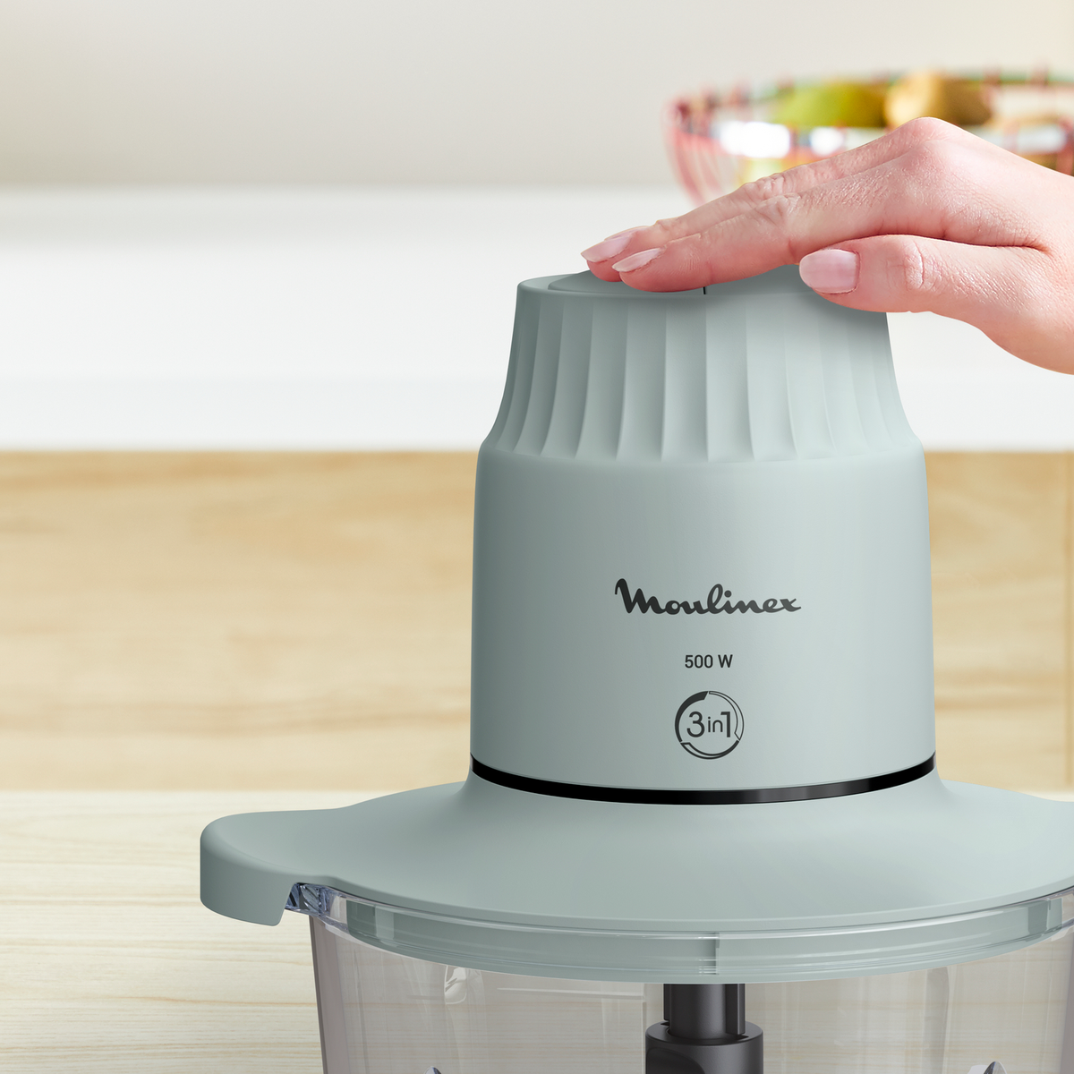 Una mano descansa sobre un procesador de alimentos Moulinex azul claro. El fondo es una cocina borrosa.