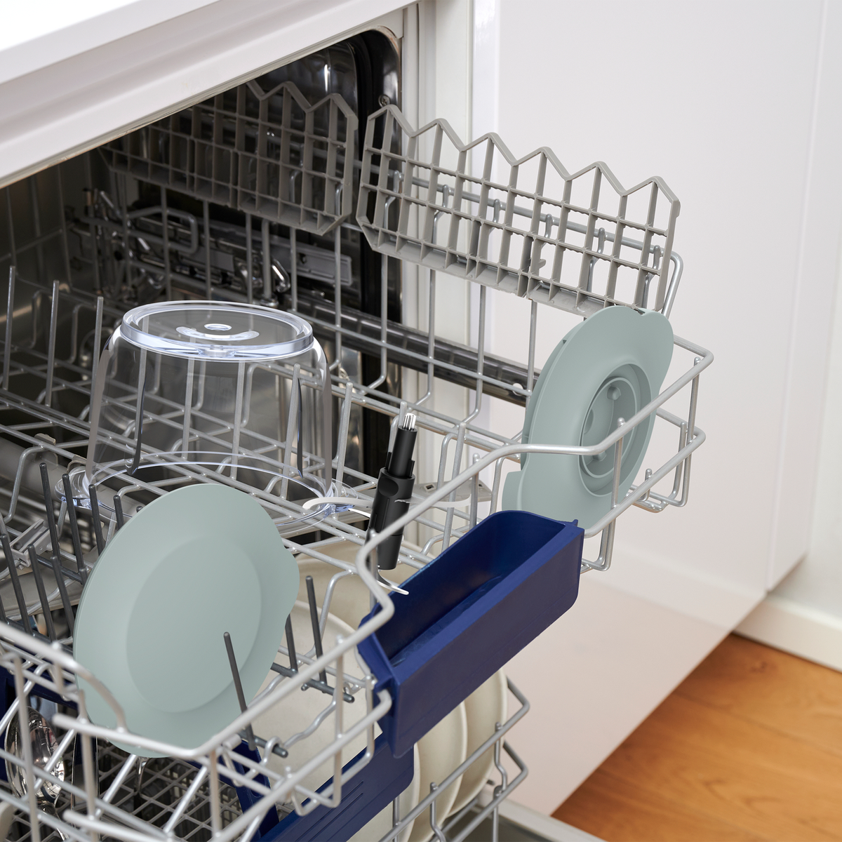 Lavavajillas abierto con platos. Platos, tazas y utensilios están en los estantes. Suelo de madera visible.