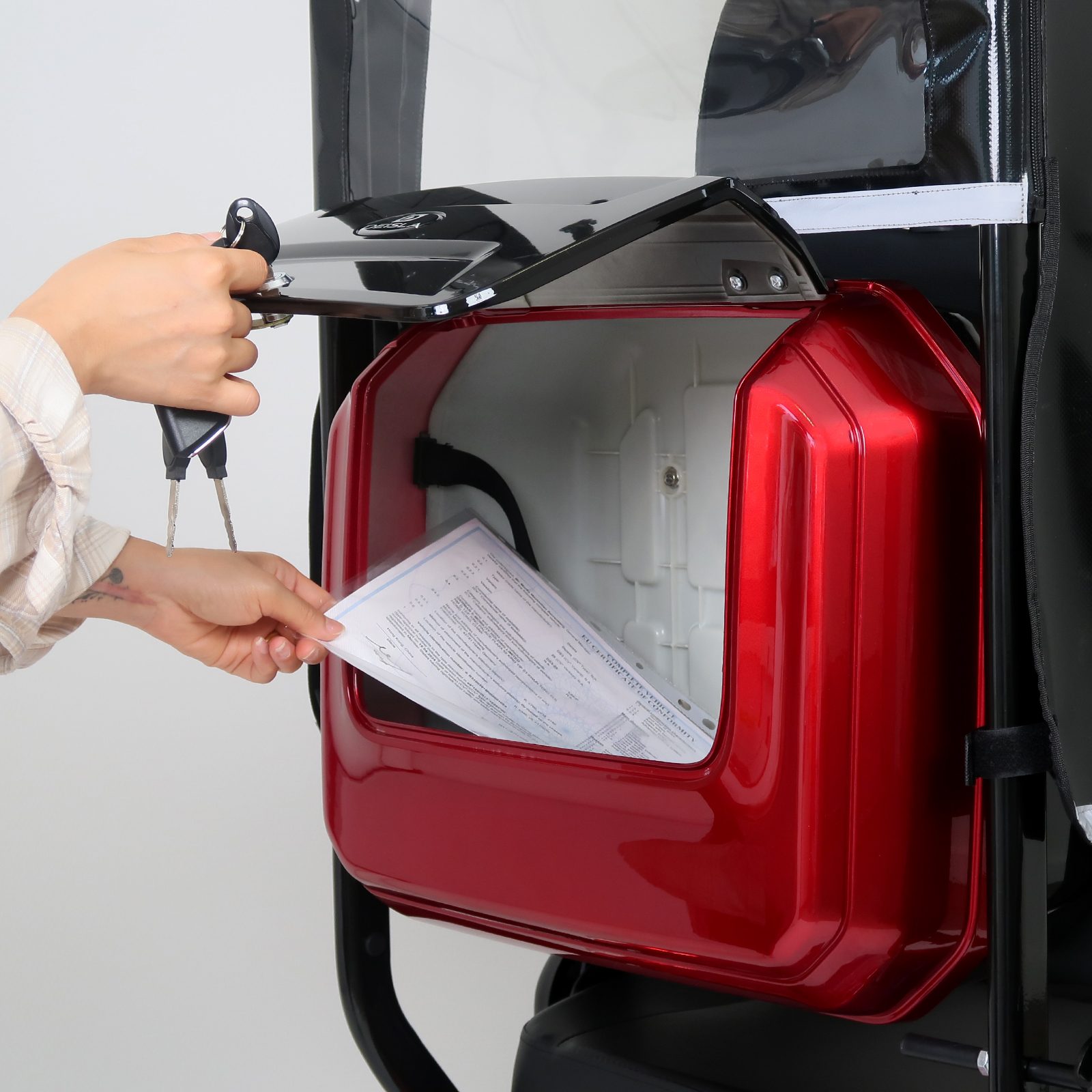 Hände öffnen ein rotes Staufach an einem roten Mobilitätsroller. Sie halten ein Papier.