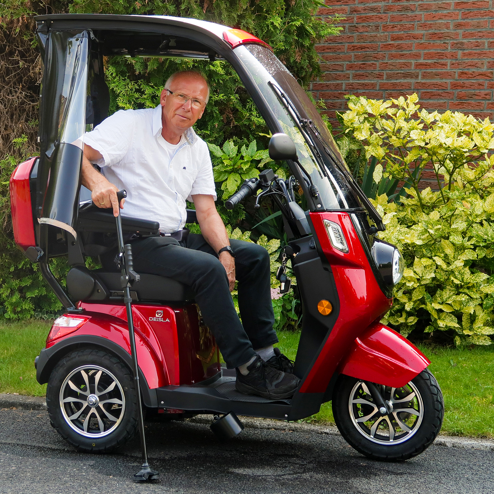 Ein Mann in weißem Hemd und schwarzer Hose sitzt auf einem roten Mobilitätsroller mit Verdeck. Im Freien.