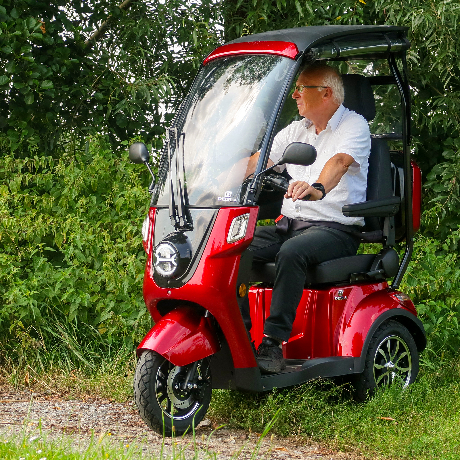 Ein älterer Mann in einem weißen Hemd sitzt auf einem roten Mobilitätsroller und fährt auf einem Feldweg neben Grün.