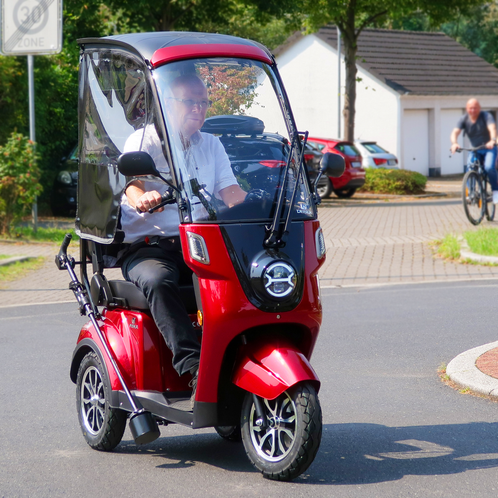 Mann fährt mit einem roten Roller mit Verdeck auf einer Straße. Eine andere Person auf einem Fahrrad ist im Hintergrund sichtbar.