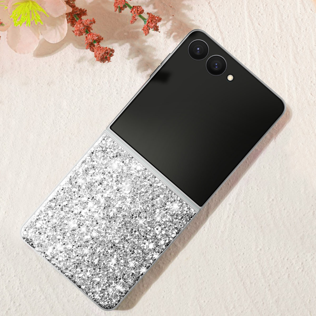 Coque de téléphone argentée à paillettes. Écran et caméra noirs. Fleurs roses en arrière-plan.