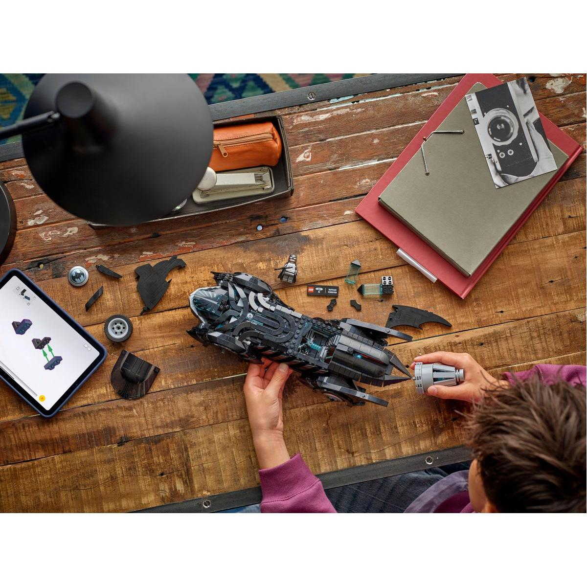 Una persona che assembla un Batmobile Lego nero su un tavolo di legno con altri pezzi Lego.