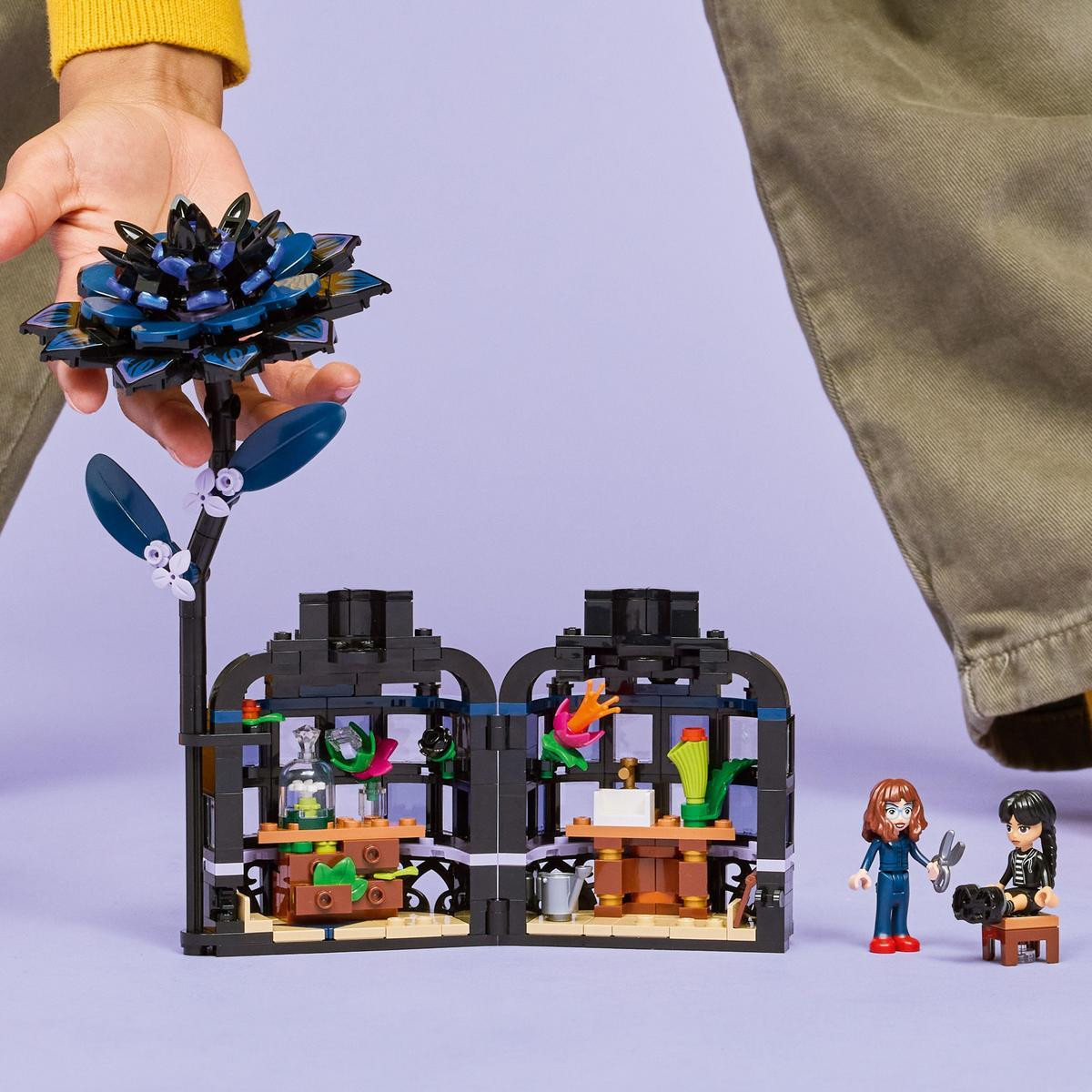 Dłoń trzymająca kwiat LEGO nad zestawem LEGO z dwoma pokojami i dwiema minifigurkami.