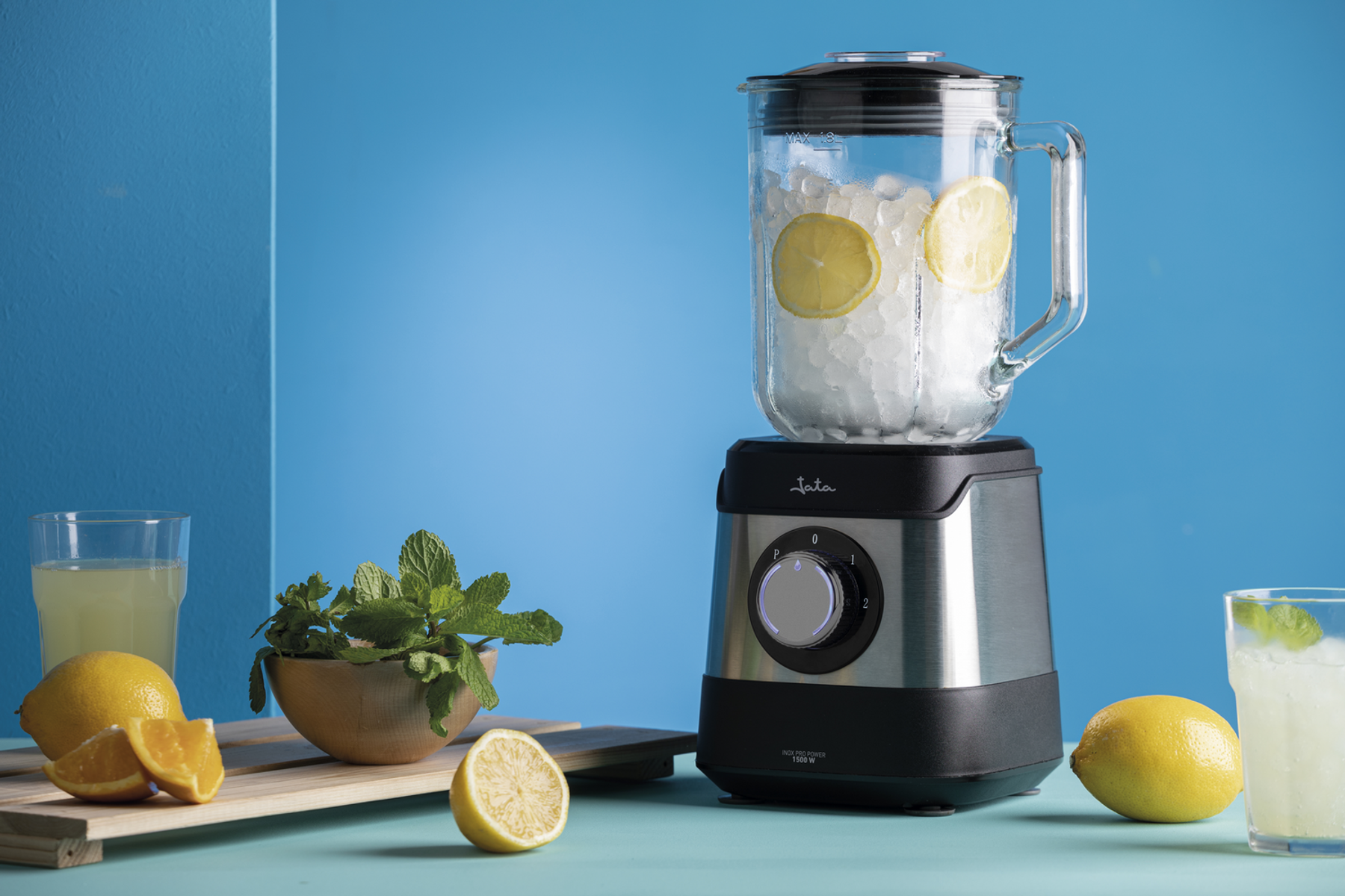 Un frullatore con ghiaccio, fettine di limone e menta all'interno. Limoni e altri ingredienti nelle vicinanze.