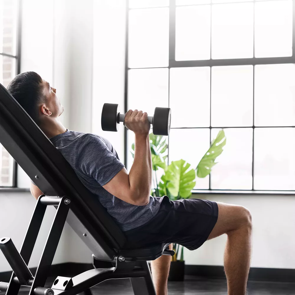 A man doing a dumbbell chest exercise on an adjustable bench near a window.