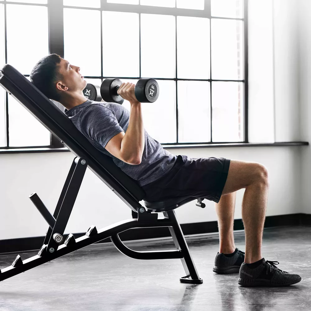 A man doing a dumbbell chest exercise on an adjustable bench in front of a window.