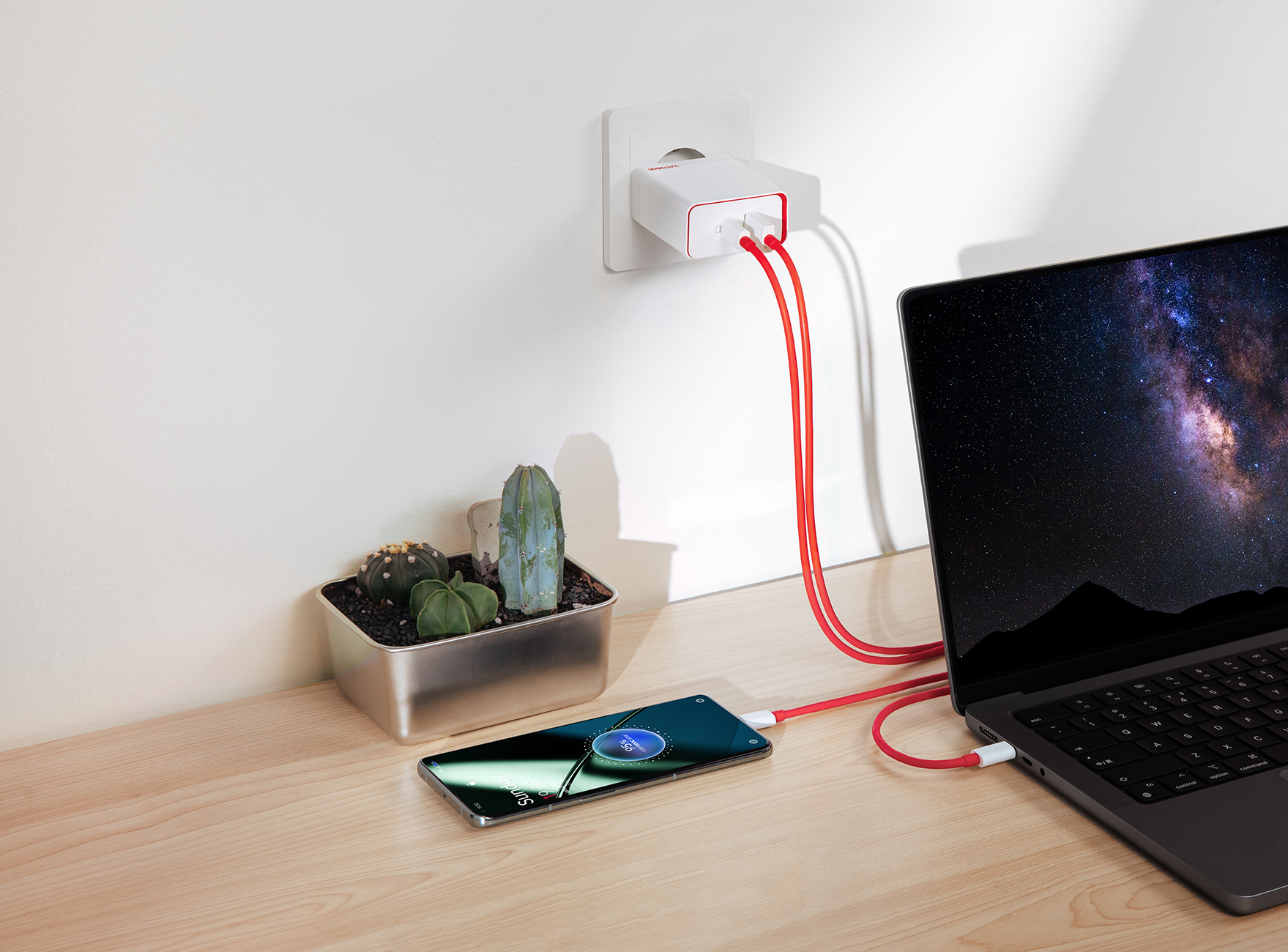Laptop, telefon i ładowarka są na biurku. Obecna jest również roślina.