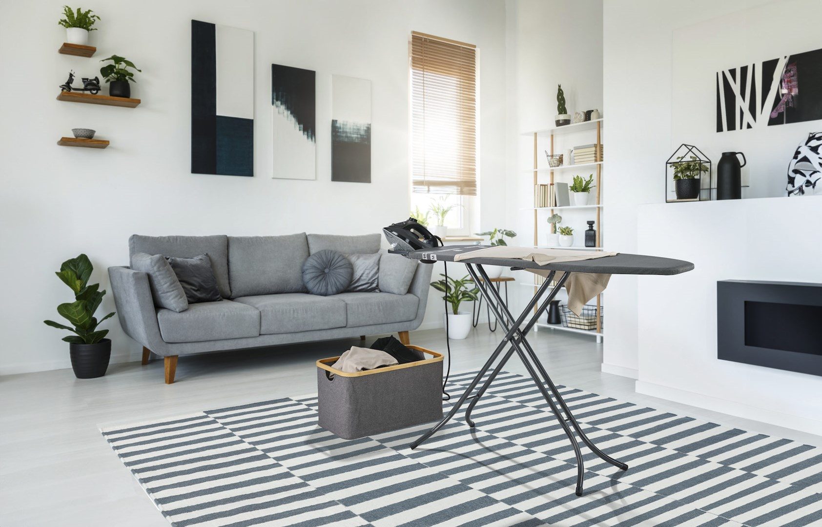Scena w salonie z szarą sofą, dywanem i rozłożoną deską do prasowania.