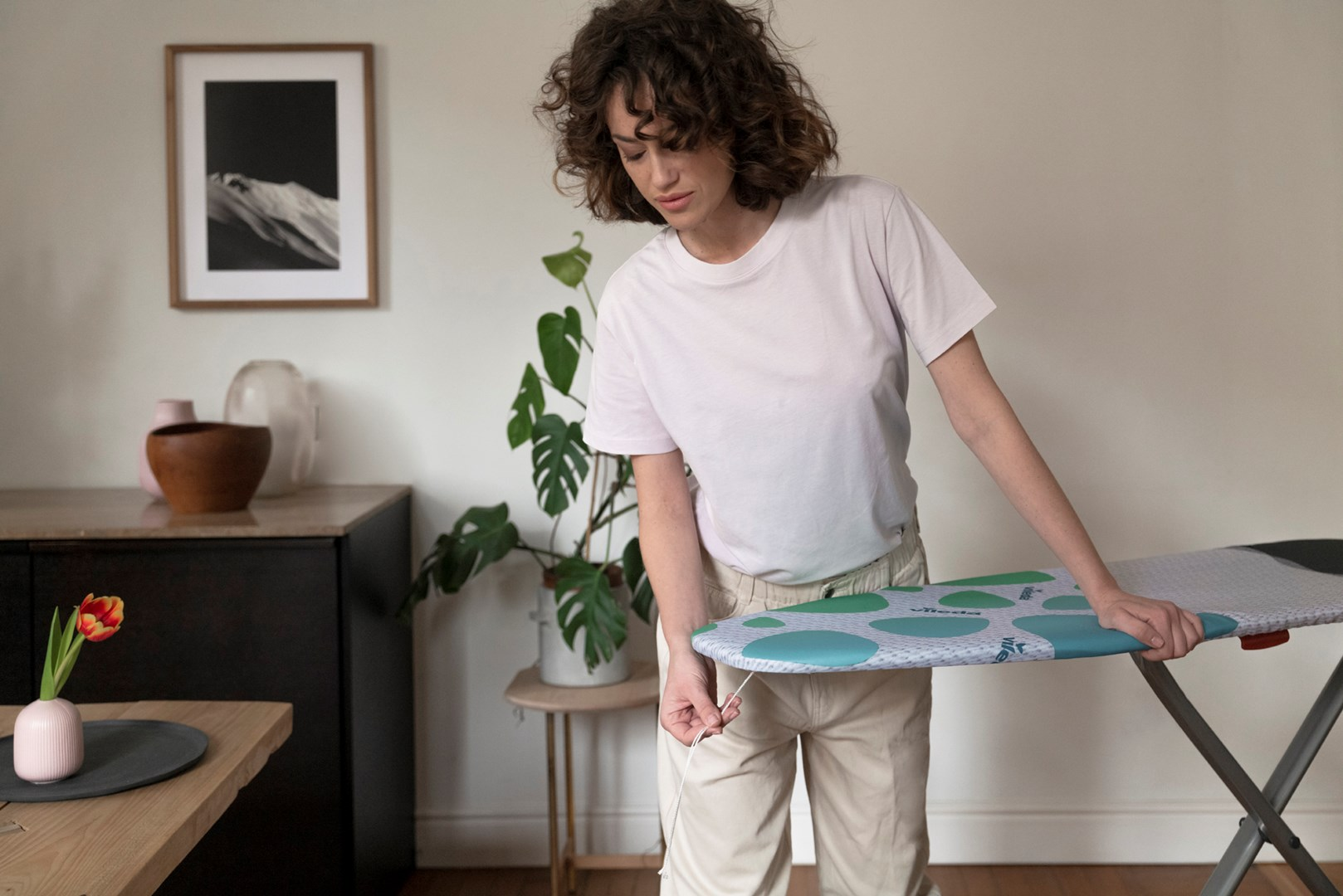 Kobieta prasuje na desce do prasowania. Ma na sobie jasnoróżową koszulę i beżowe spodnie. Pokój z drewnianymi meblami.