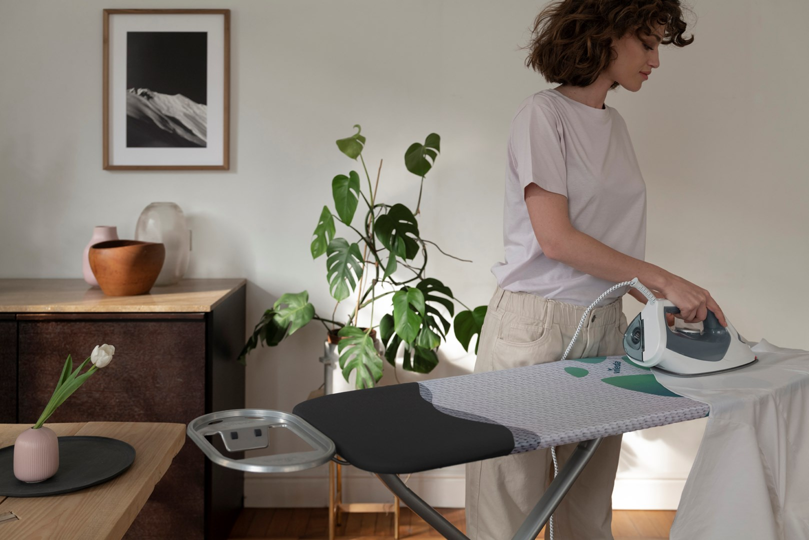 Kobieta prasuje białą tkaninę na desce do prasowania. Roślina doniczkowa w tle.