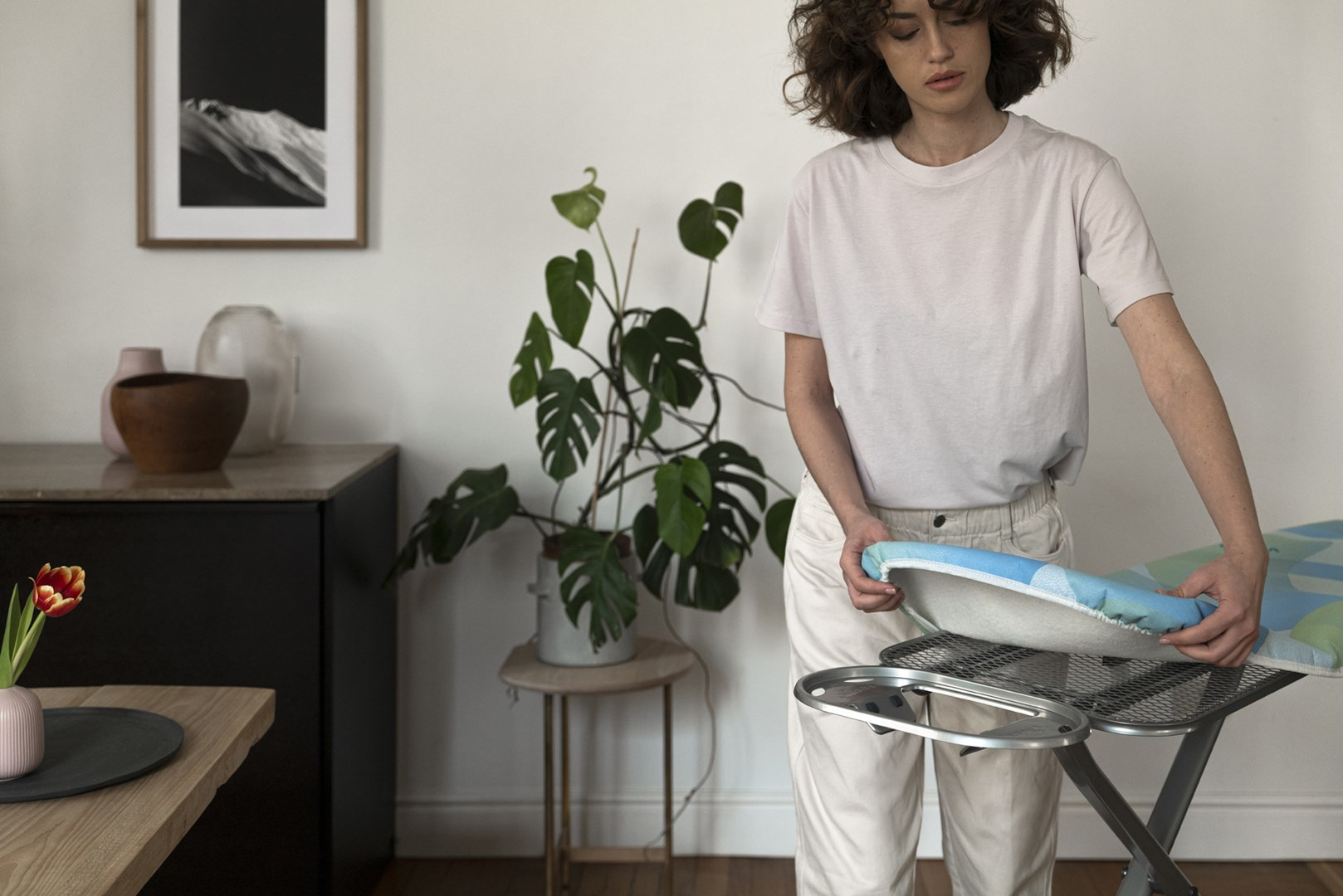 Kobieta reguluje przedłużenie deski do prasowania. Roślina doniczkowa jest widoczna w tle.