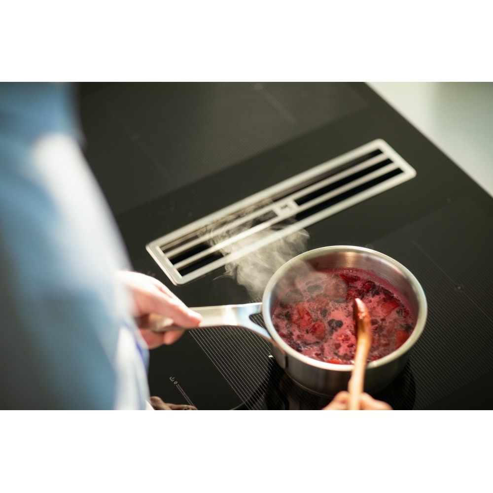 Mani che tengono una pentola con marmellata bollente. La pentola è su un piano cottura nero con una cappa.