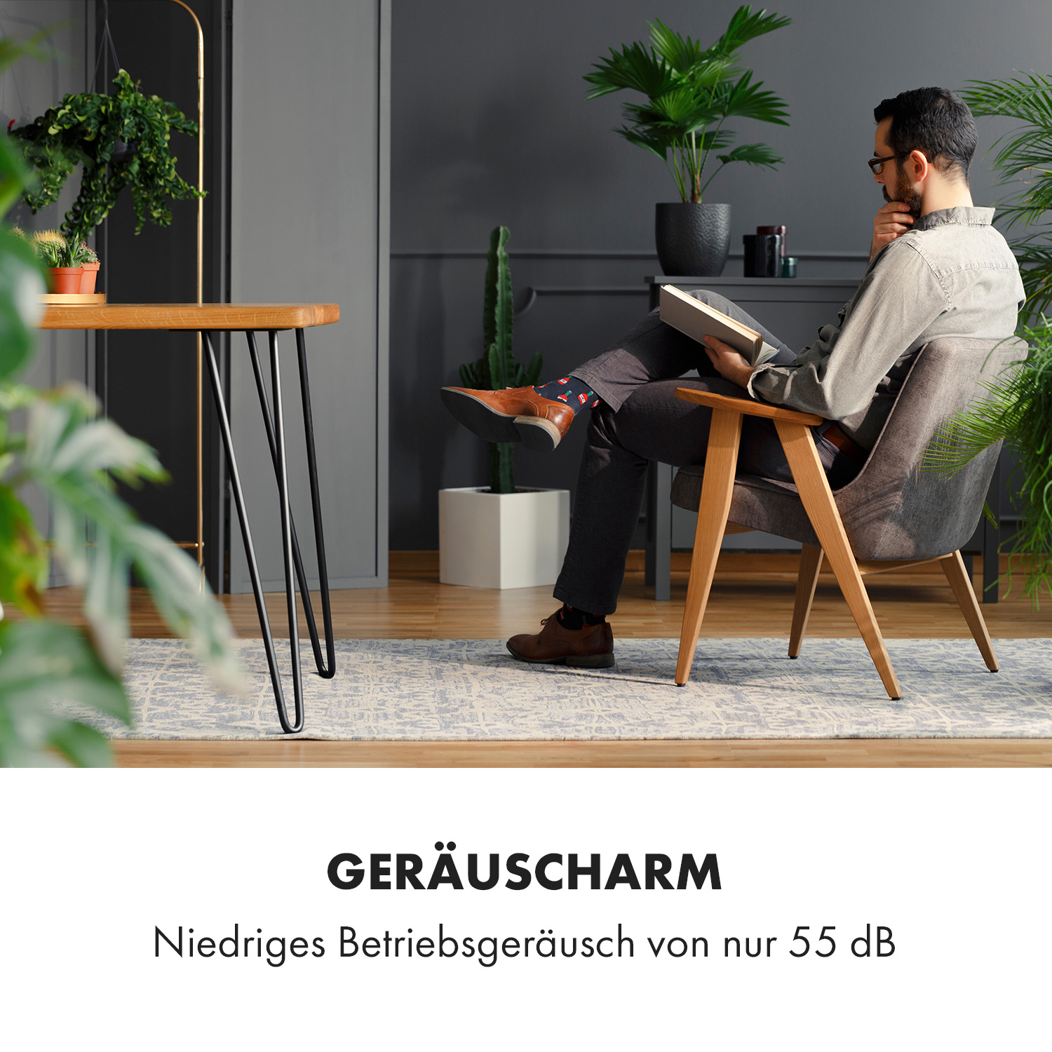 Ein Mann sitzt in einem grauen Stuhl und liest ein Buch. Es gibt einen Holztisch und Pflanzen um ihn herum. Der Text lautet "GERÄUSCHARM".