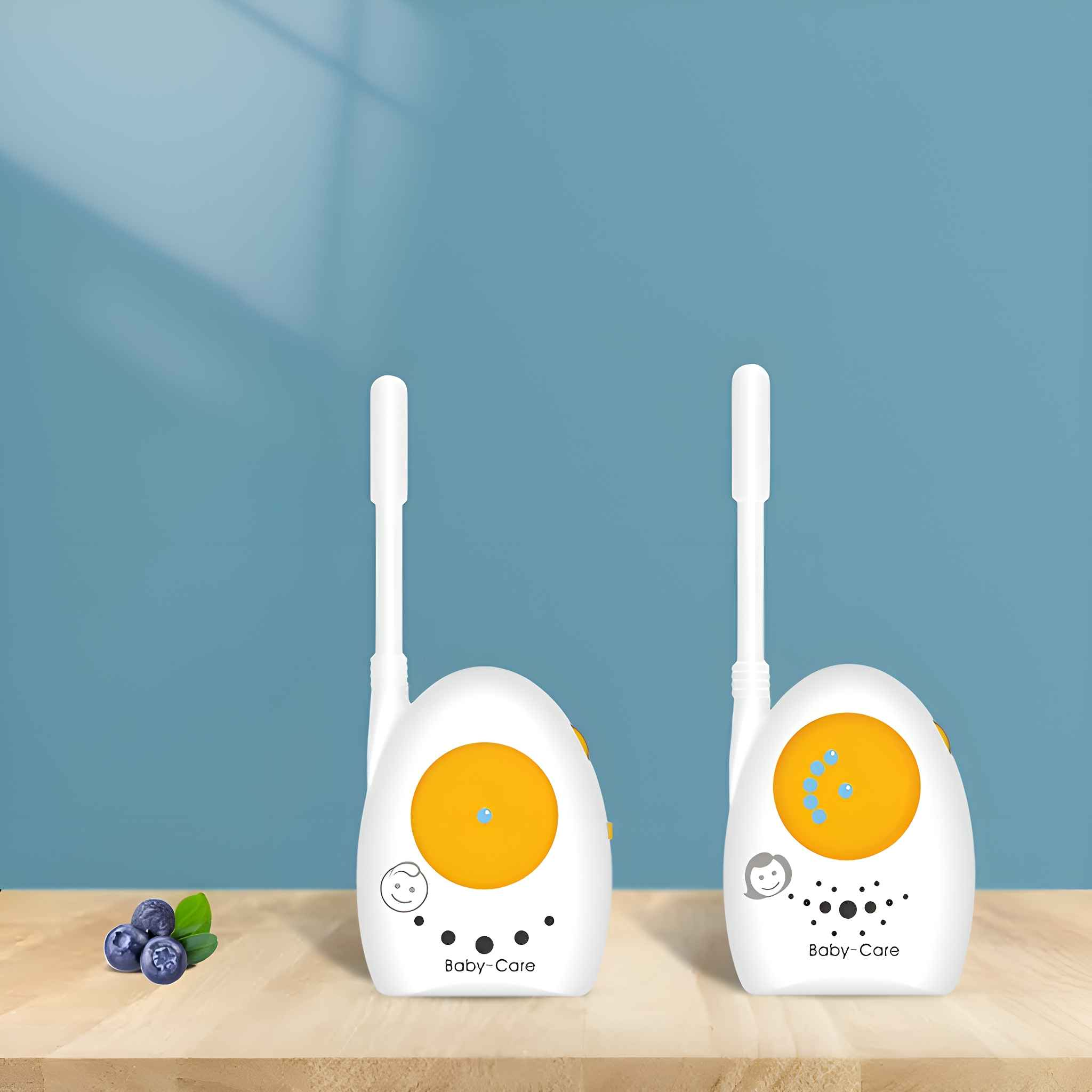 Zwei weiße Babyphone mit Antennen. Blaubeeren liegen links auf einer Holzoberfläche vor einer blauen Wand.