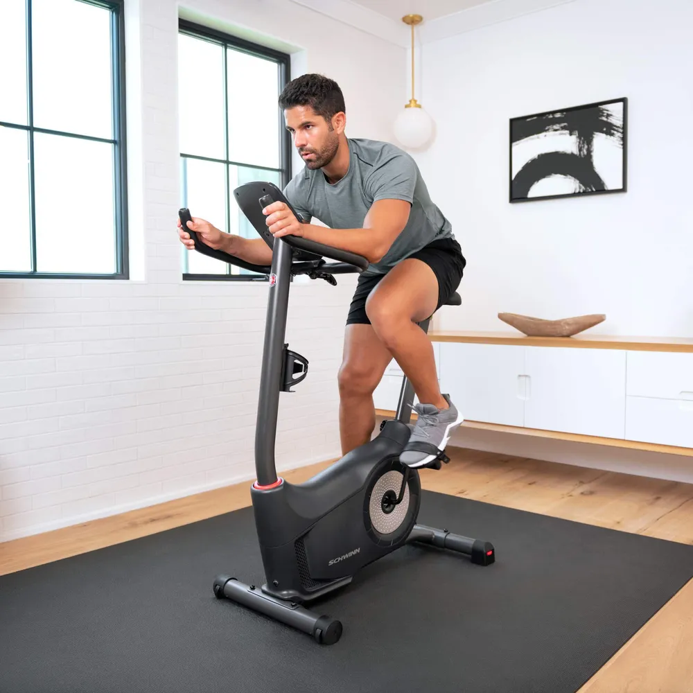 A man on a stationary bike is exercising. The bike is black and in a room with windows.