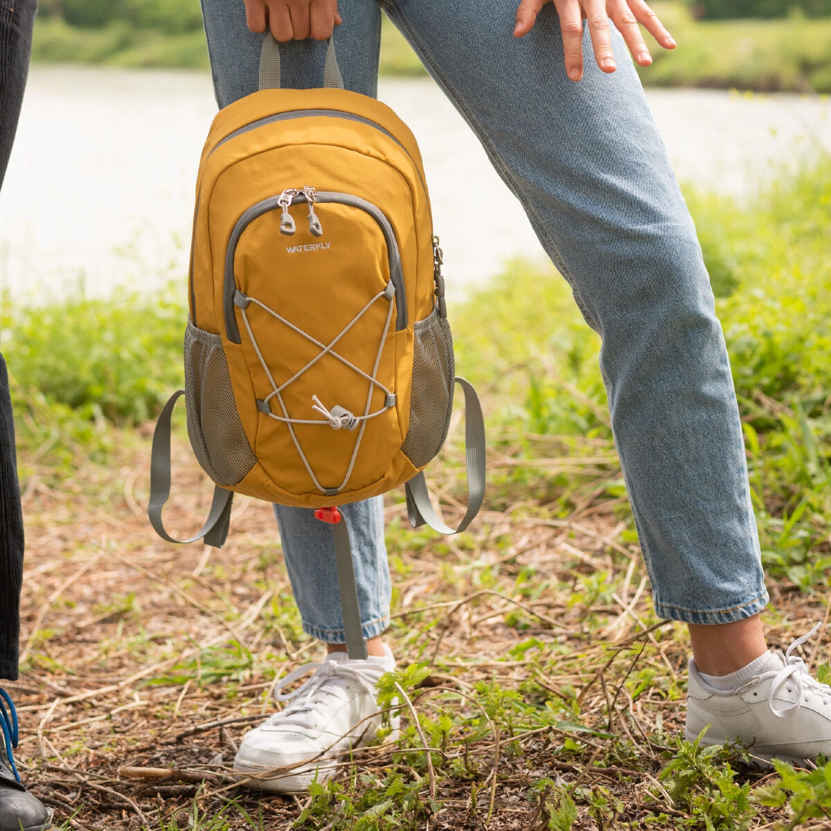 Una persona con jeans e scarpe bianche, che tiene uno zaino giallo vicino a un corso d'acqua.
