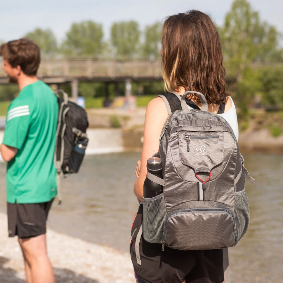 Zwei Personen mit Rucksäcken am Wasser. Eine Person macht ein Foto mit ihrem Handy.