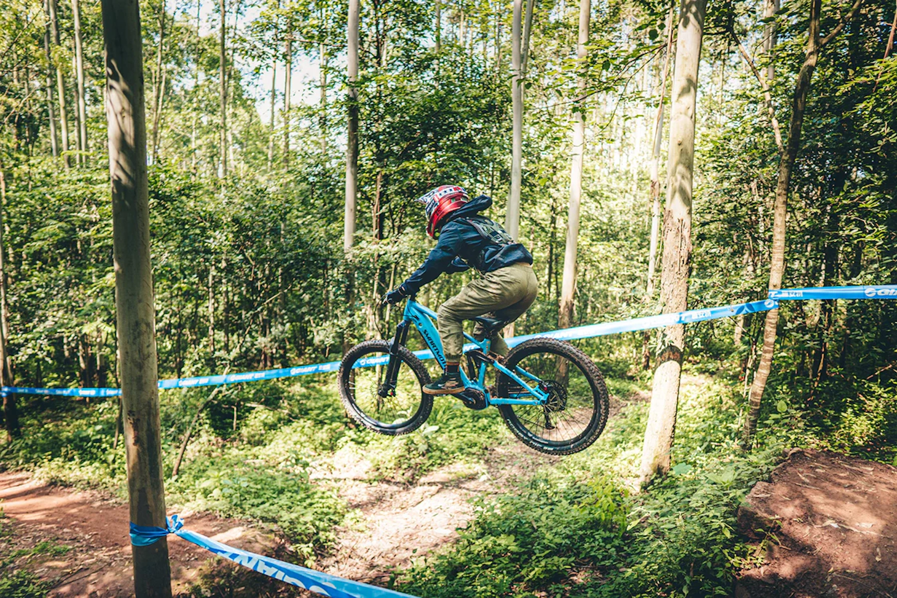 Ein Mountainbiker springt über ein blaues Band auf einem blauen Fahrrad. Waldhintergrund mit Bäumen und Grün.