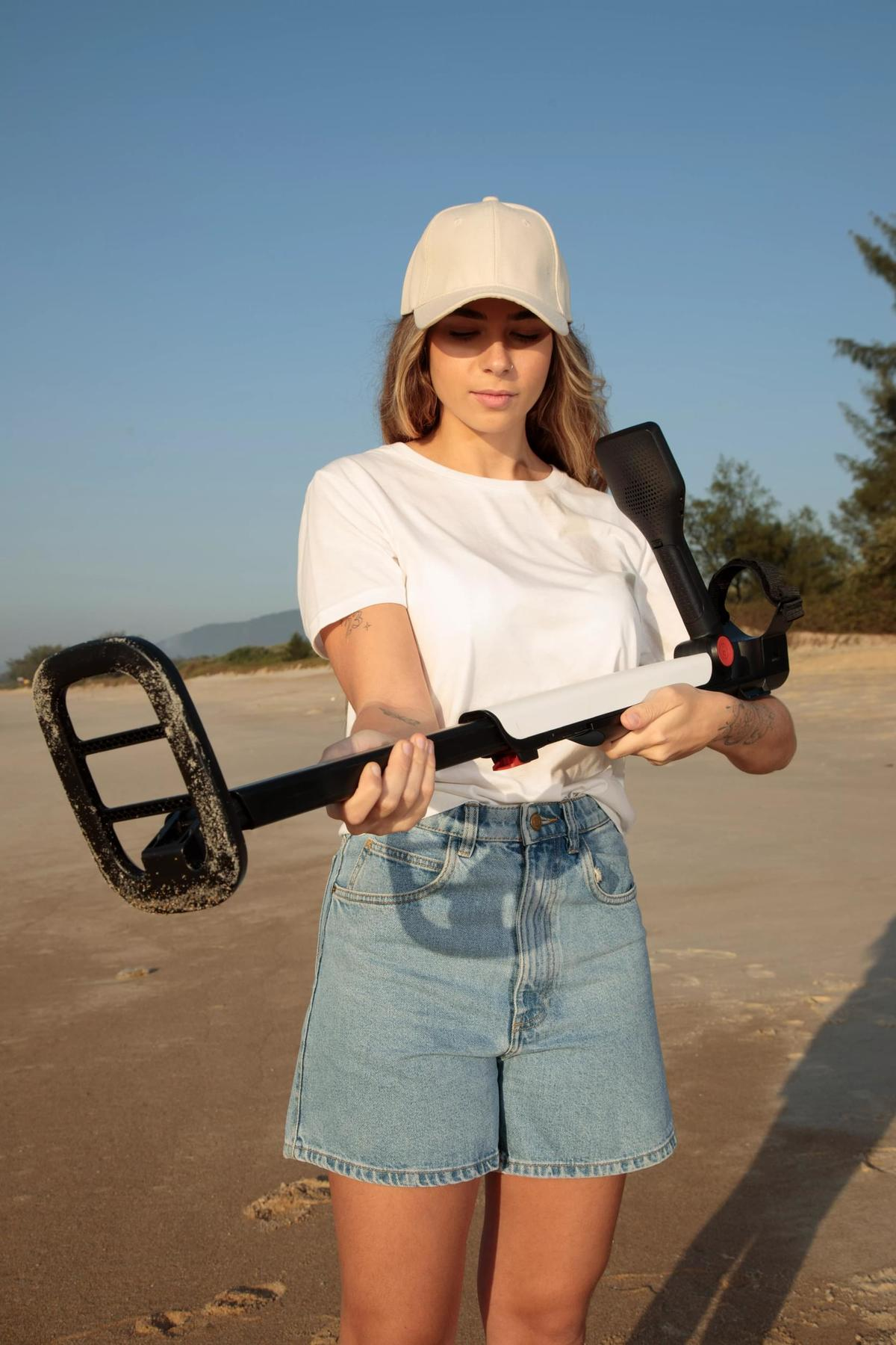 Una donna su una spiaggia tiene in mano un metal detector. Indossa un berretto e una maglietta bianchi, e pantaloncini di jeans.