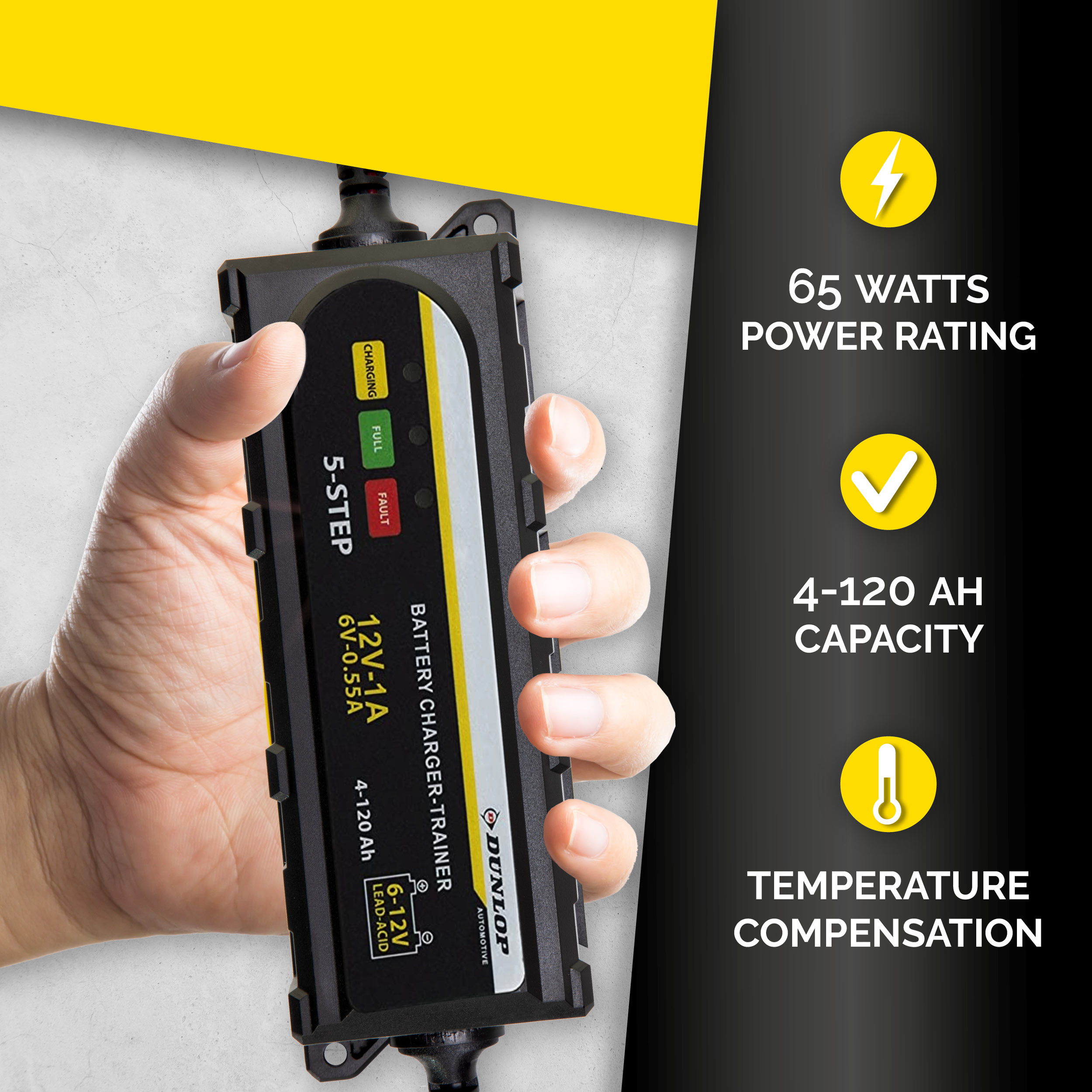 A hand holds a black battery charger with a yellow label and text. The charger has lights.