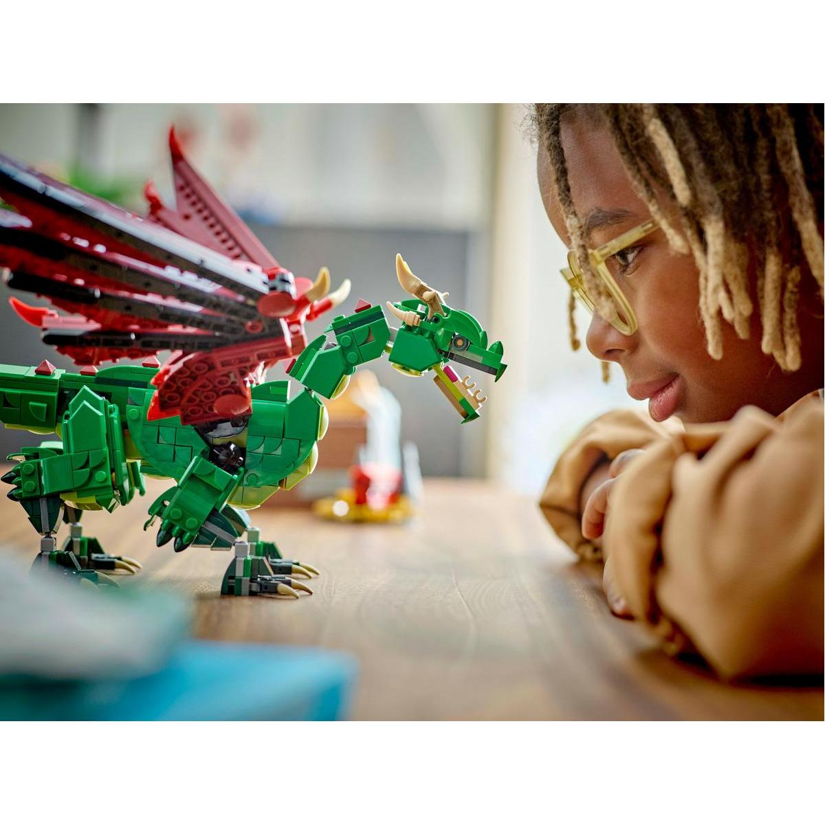 Dziecko patrzące na smoka Lego, zielonego z czerwonymi skrzydłami, umieszczonego na drewnianym stole.