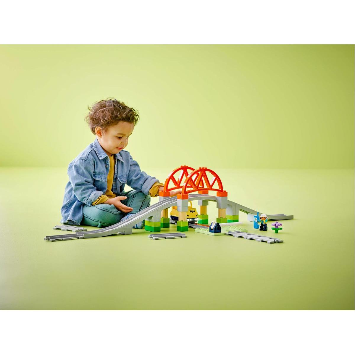 Małe dziecko bawi się zestawem kolejowym Lego Duplo, w komplecie z torami, mostem i figurkami.