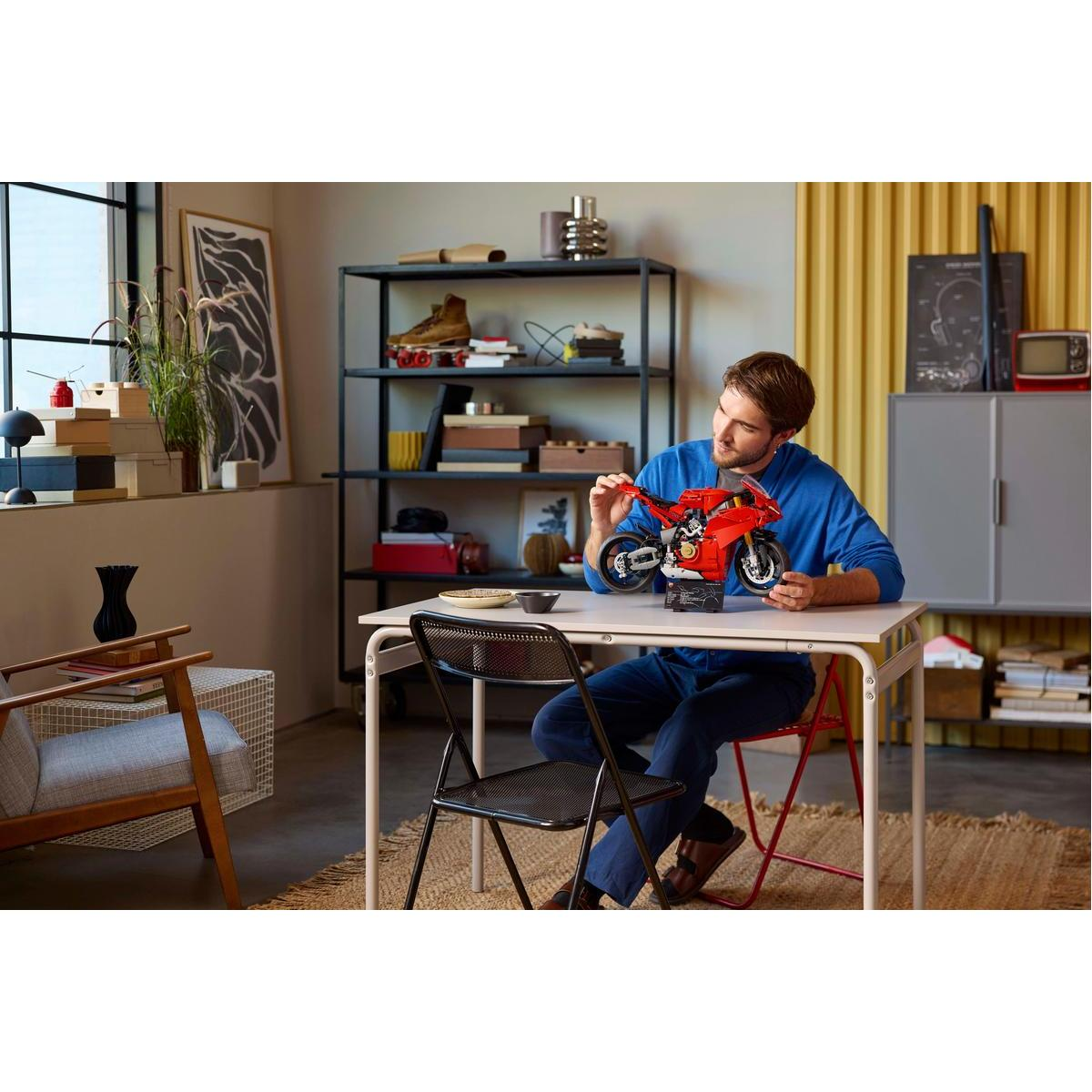 Mężczyzna siedzi przy stole, składając czerwony zestaw Lego motocykla. Scena obejmuje okno, półki i czarne krzesło.