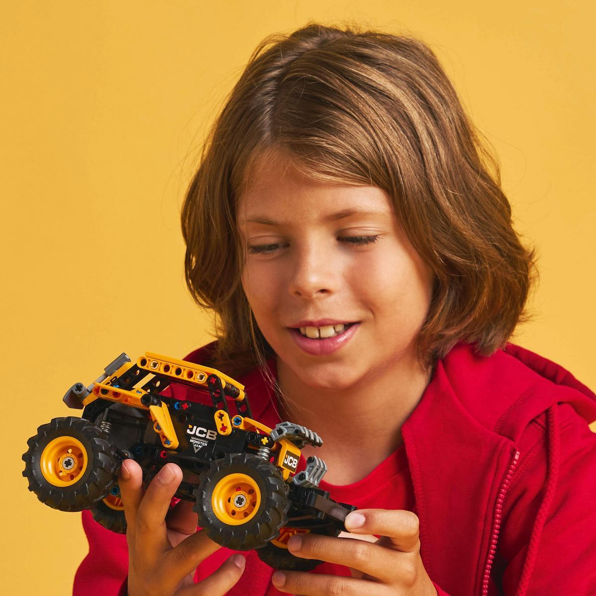 Un niño sonríe mientras mira un camión monstruo Lego. El camión es amarillo y negro y tiene ruedas grandes.