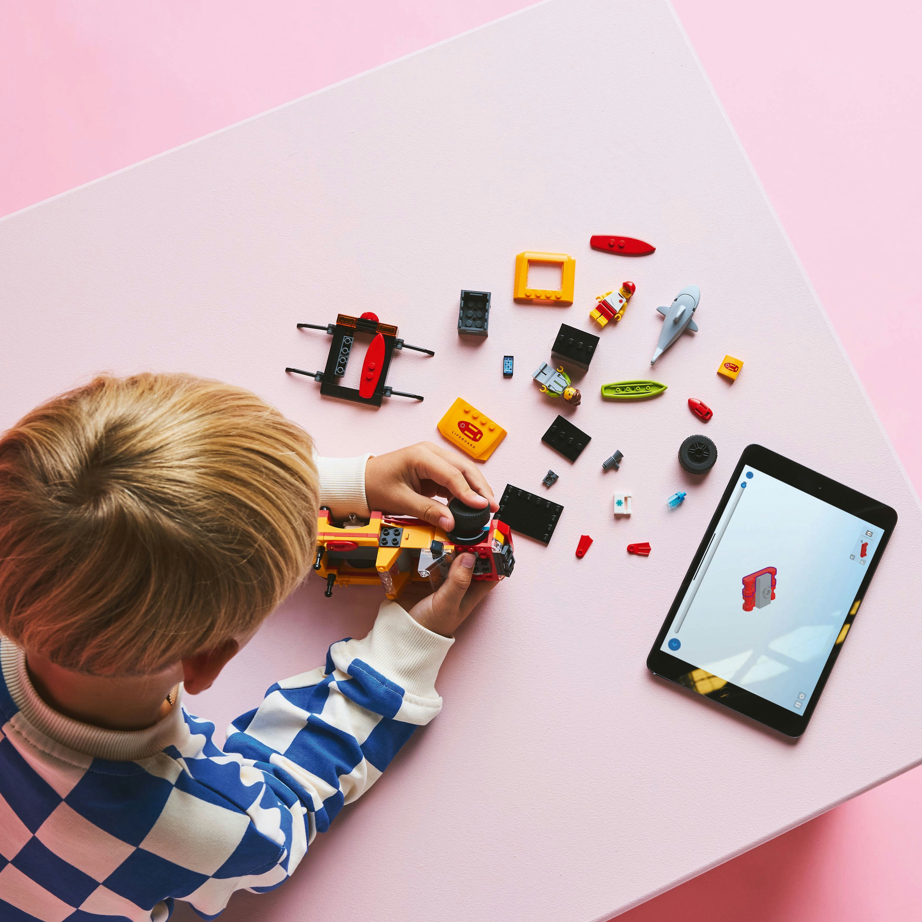 Un enfant joue avec des pièces Lego et une tablette sur une surface rose.