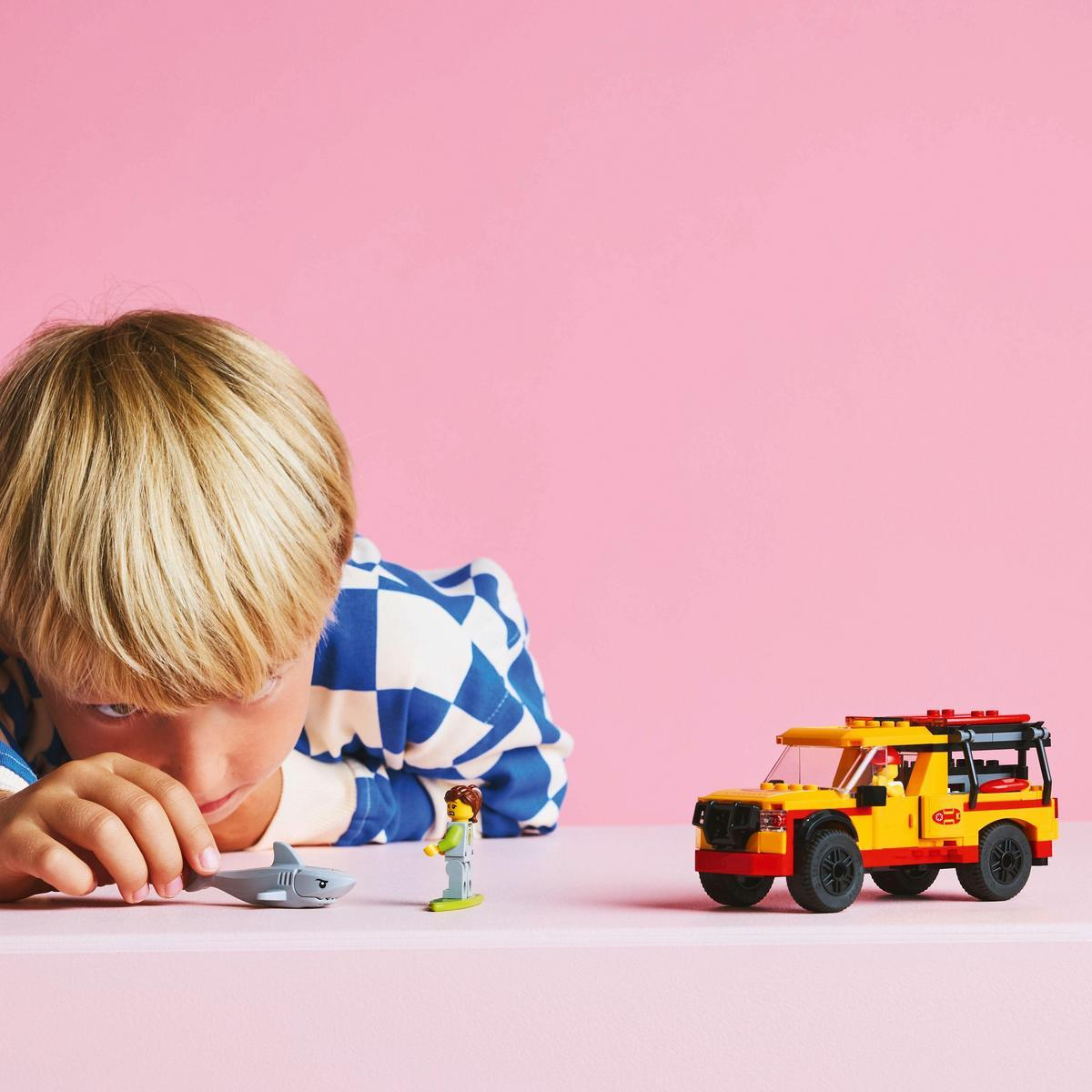 Un garçon regarde un requin jouet et un set de voiture Lego sur une surface rose.