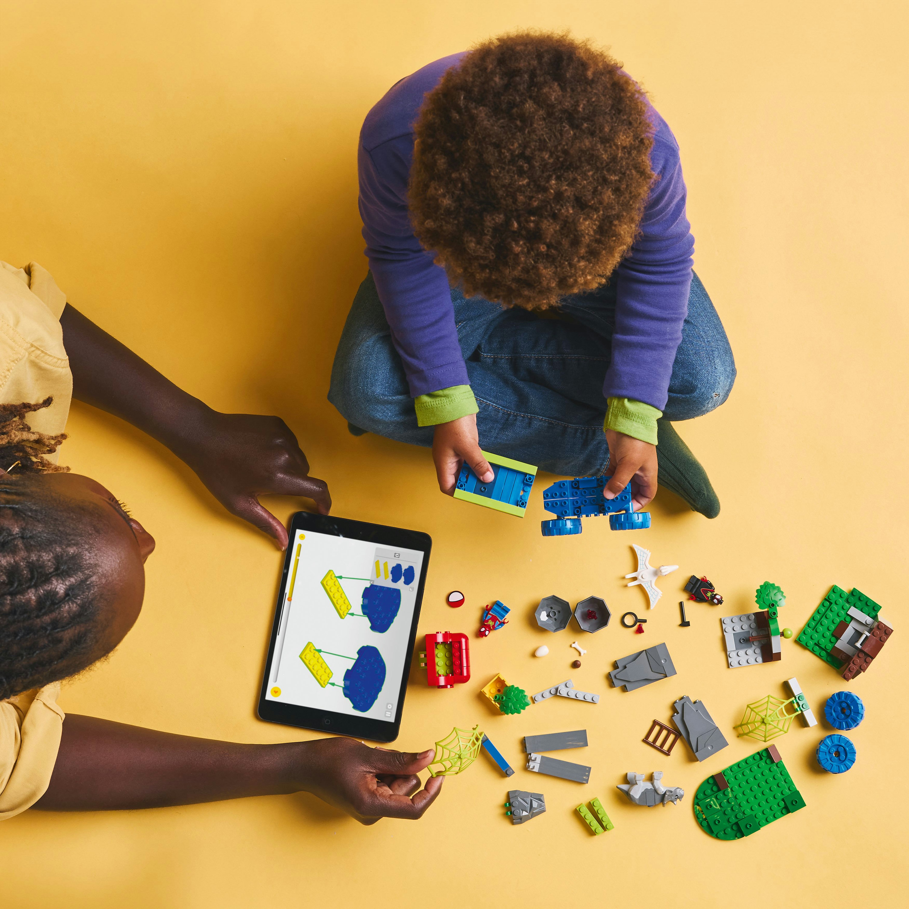 Zwei Personen mit einem Tablet und Lego-Teilen auf gelbem Hintergrund.