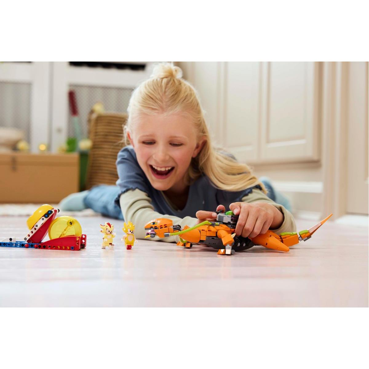 Dziewczynka leży na podłodze, bawiąc się pomarańczowym smokiem Lego. W pobliżu są dwie małe figurki Lego, uśmiechnięte.