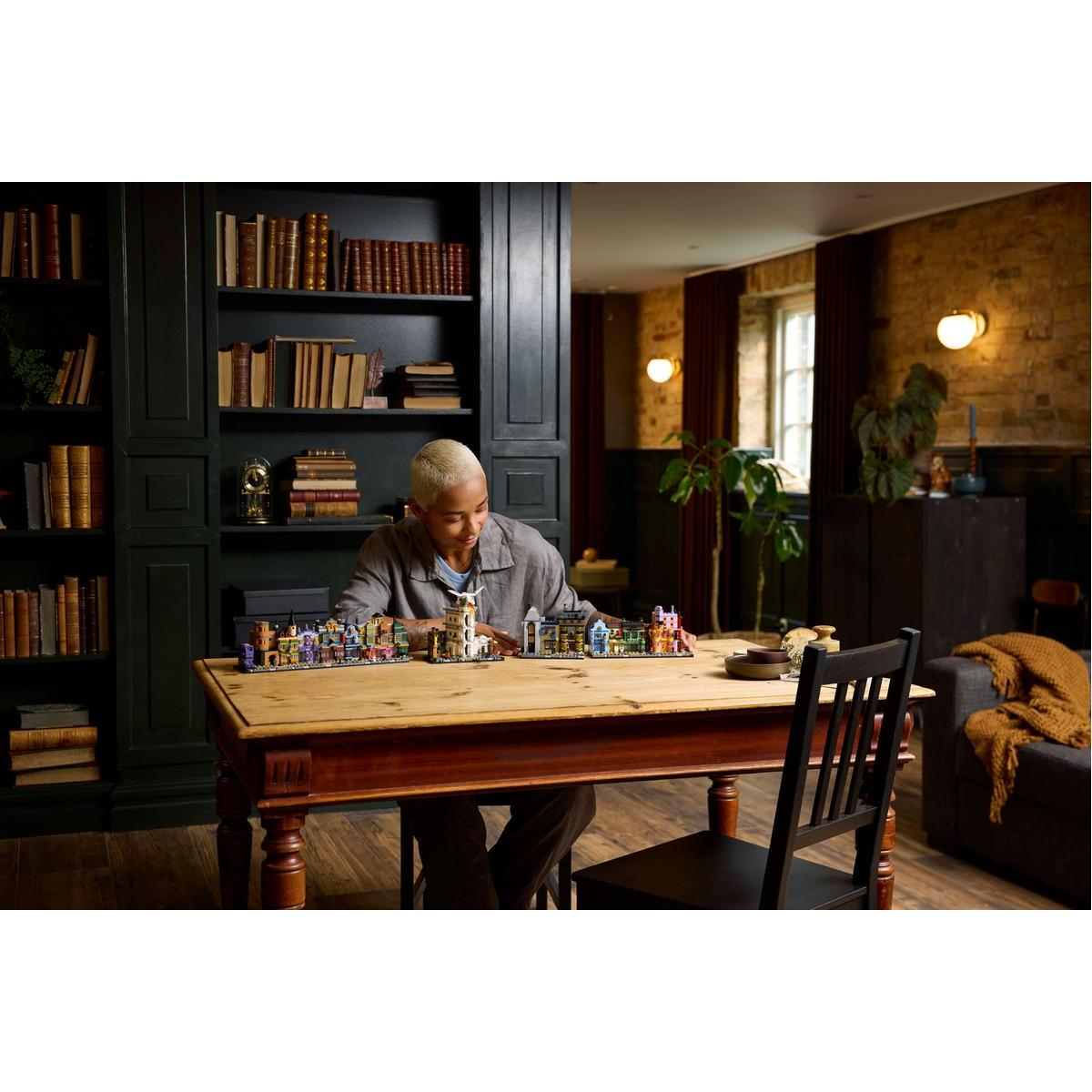 Osoba patrzy na zestaw miasta Lego na stole. Zestaw jest kolorowy, z wieloma budynkami. W pobliżu regały z książkami i kanapa.