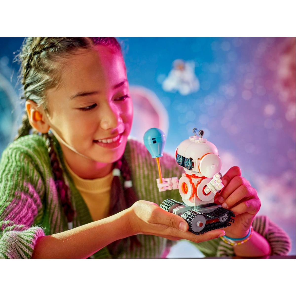 Dziewczyna trzyma robota Lego z balonem. Robot jest biało-pomarańczowy. Uśmiecha się do niego na niebieskim tle.