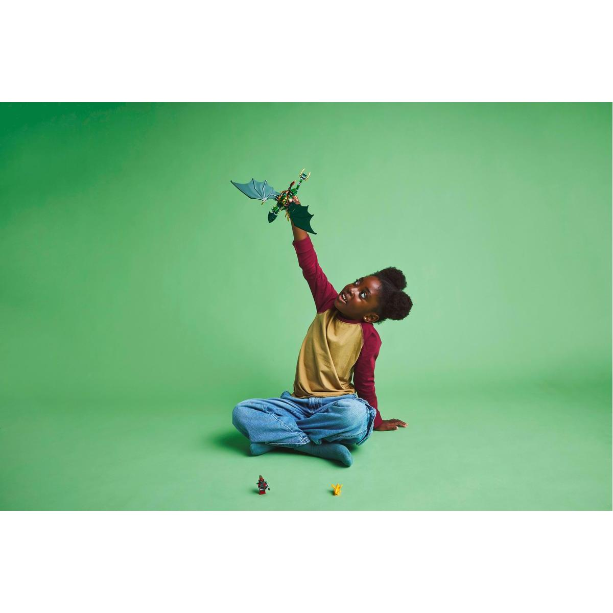 Dziewczynka siedzi na zielonym tle, trzymając smoka Lego. Smok jest zielony i złoty.