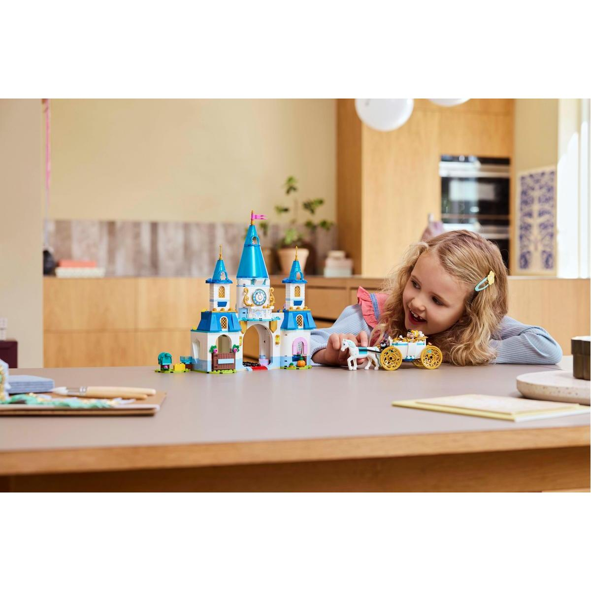 Dziewczynka bawi się zamkiem i powozem Lego na stole. Zamek ma niebieskie wieże.