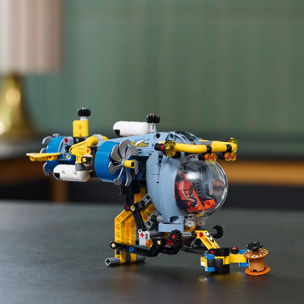 Łódź podwodna Lego na stole. Jest niebieska, żółta i biała. Wewnątrz przezroczystej kopuły znajduje się figurka.