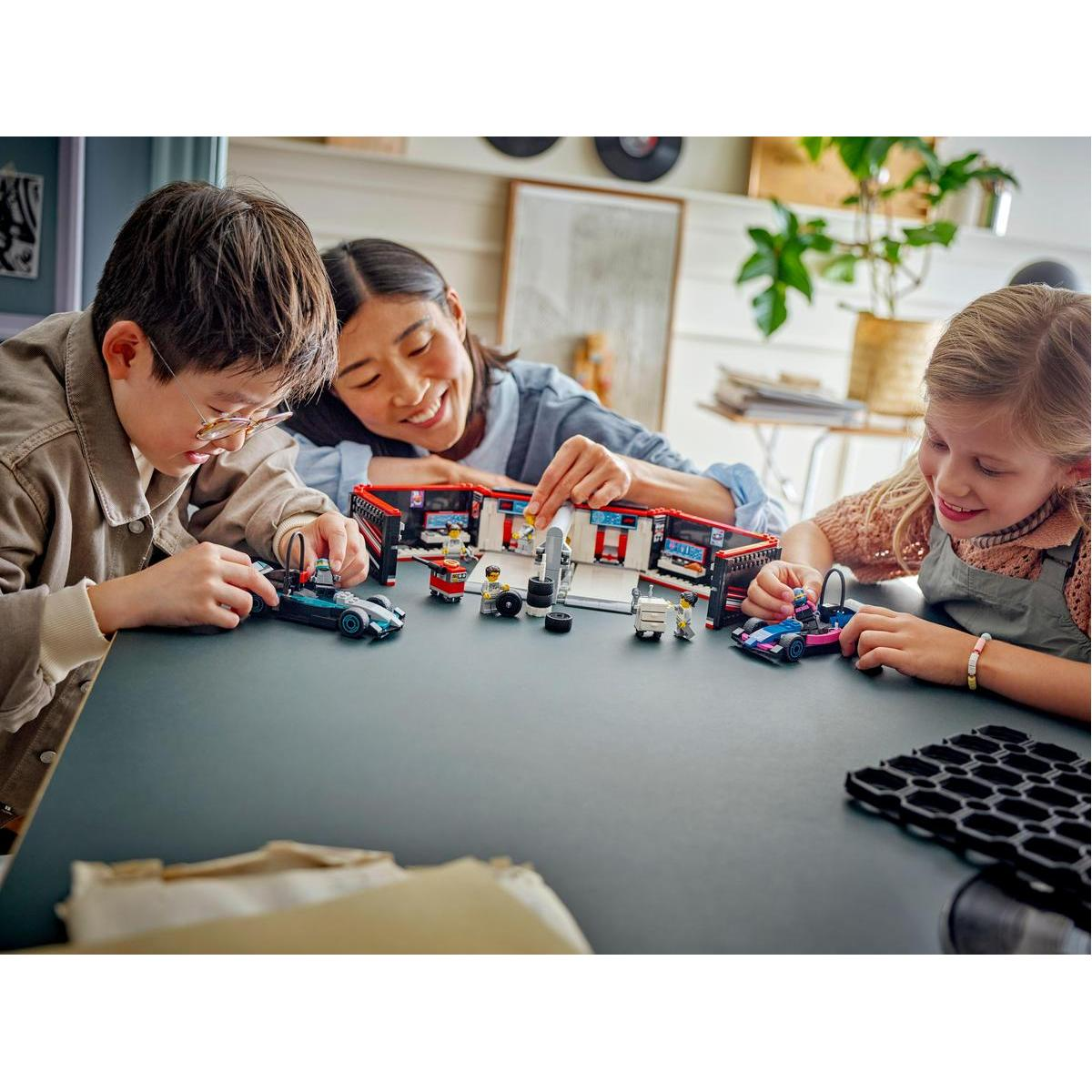 Trzy osoby bawią się samochodami Lego i zestawem garażowym na ciemnym stole. Scena jest dobrze oświetlona.